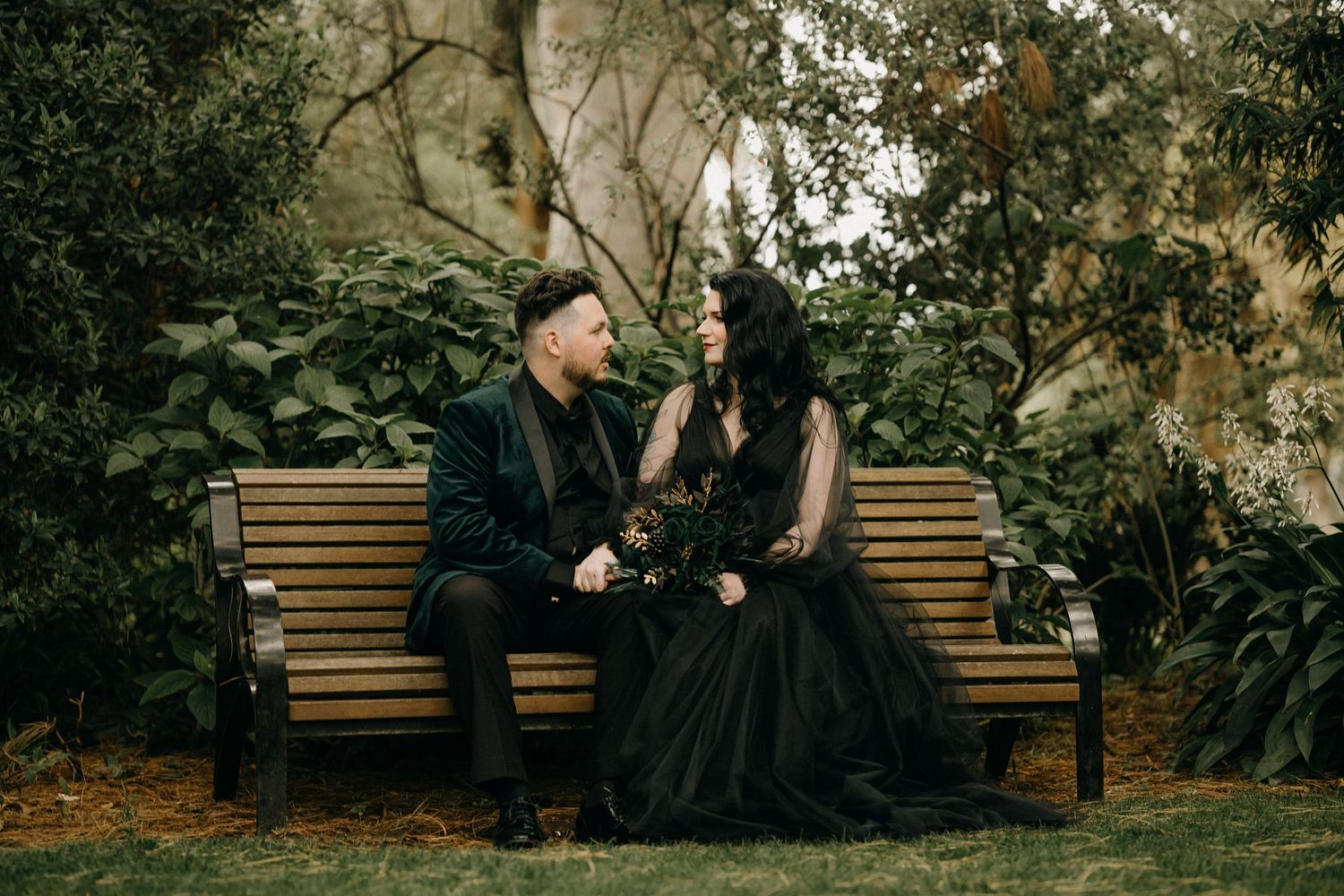 A romantic couple sits together on a wooden park bench surrounded by lush greenery and trees.