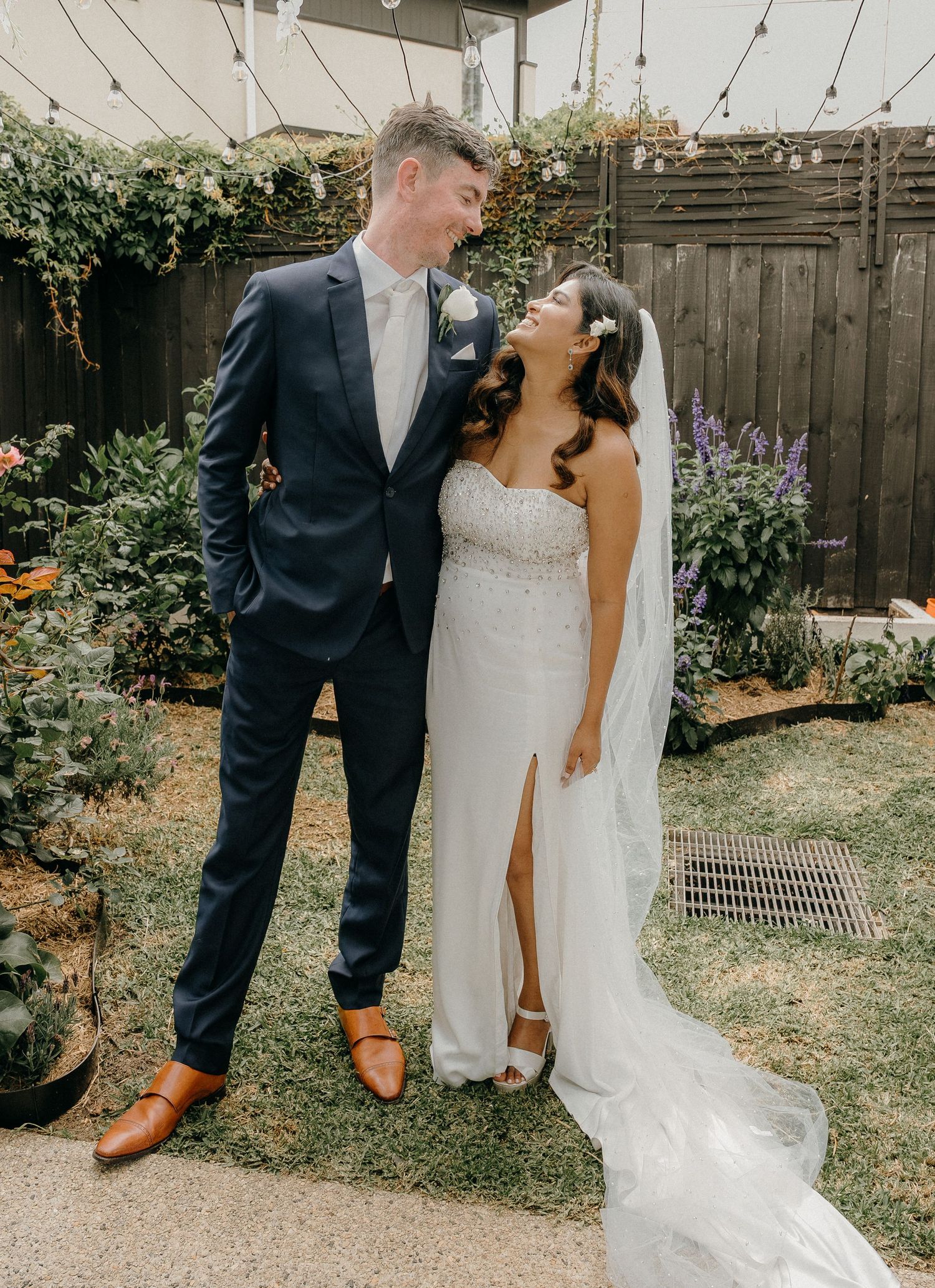 Couple in wedding attire shows off their contrasting footwear in a backyard garden setting.