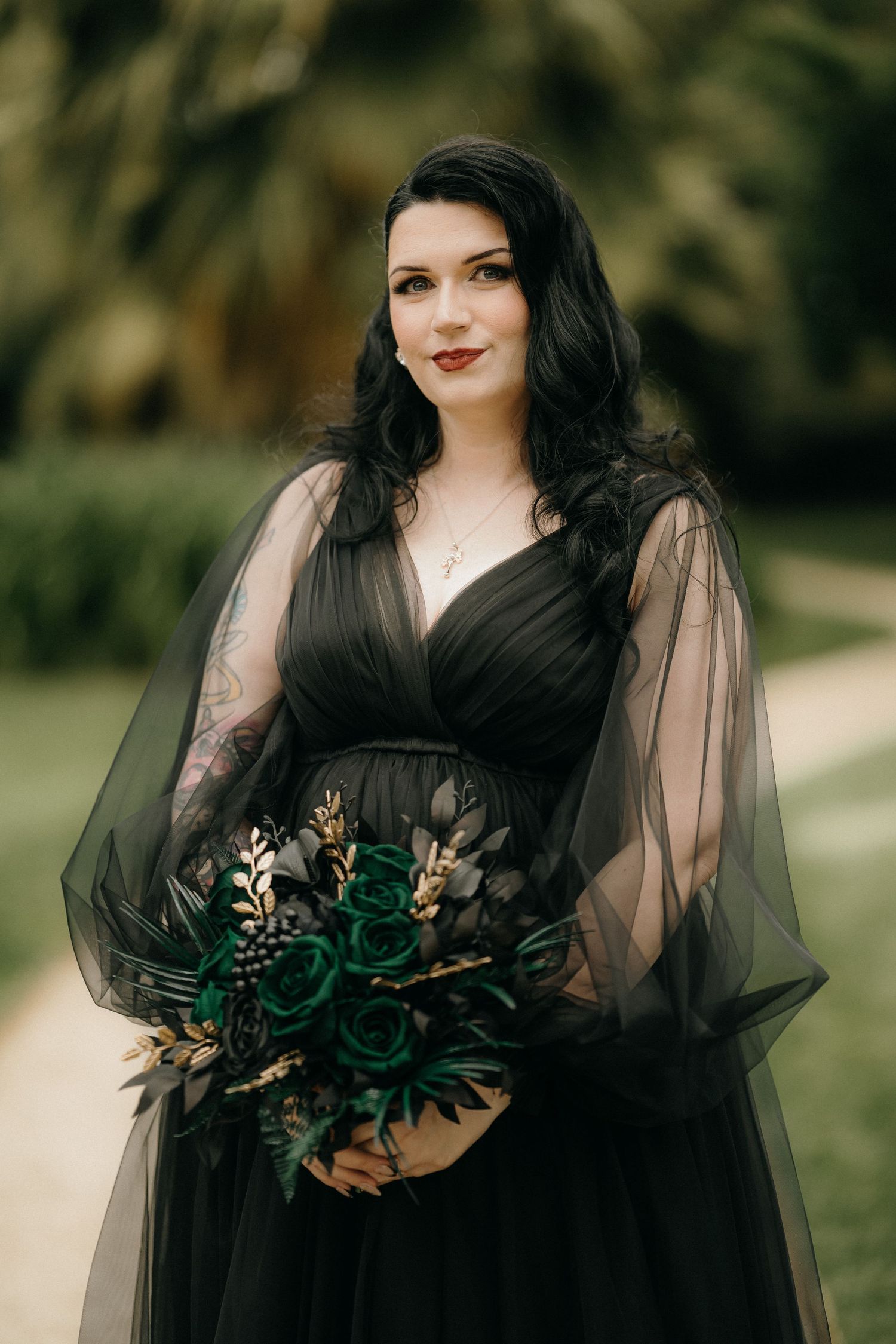 A person in a flowing black gown with sheer sleeves poses outdoors holding a dark floral bouquet.