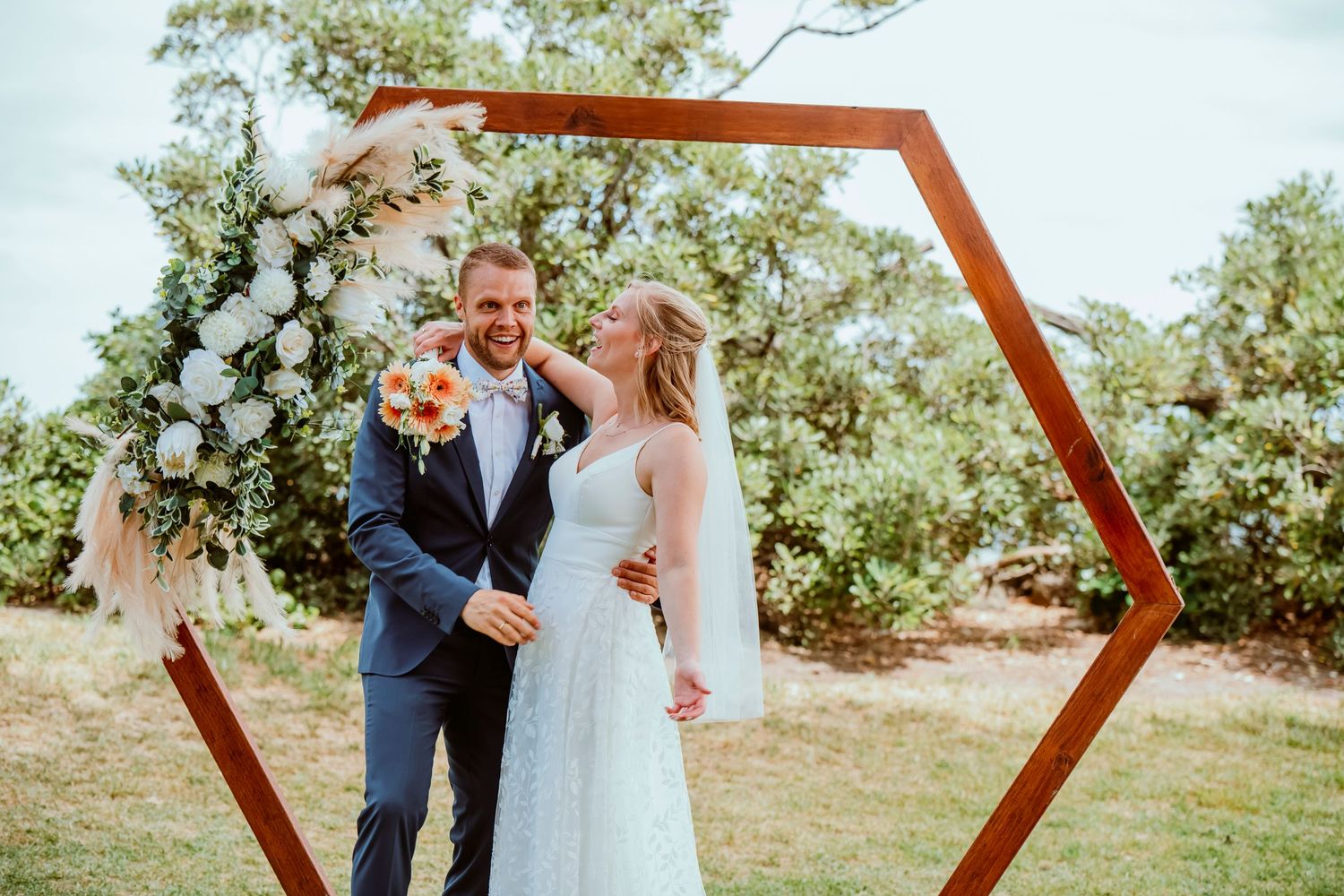 Romantic outdoor wedding ceremony with wooden hexagon arch decorated with white floral arrangements in a natural setting.