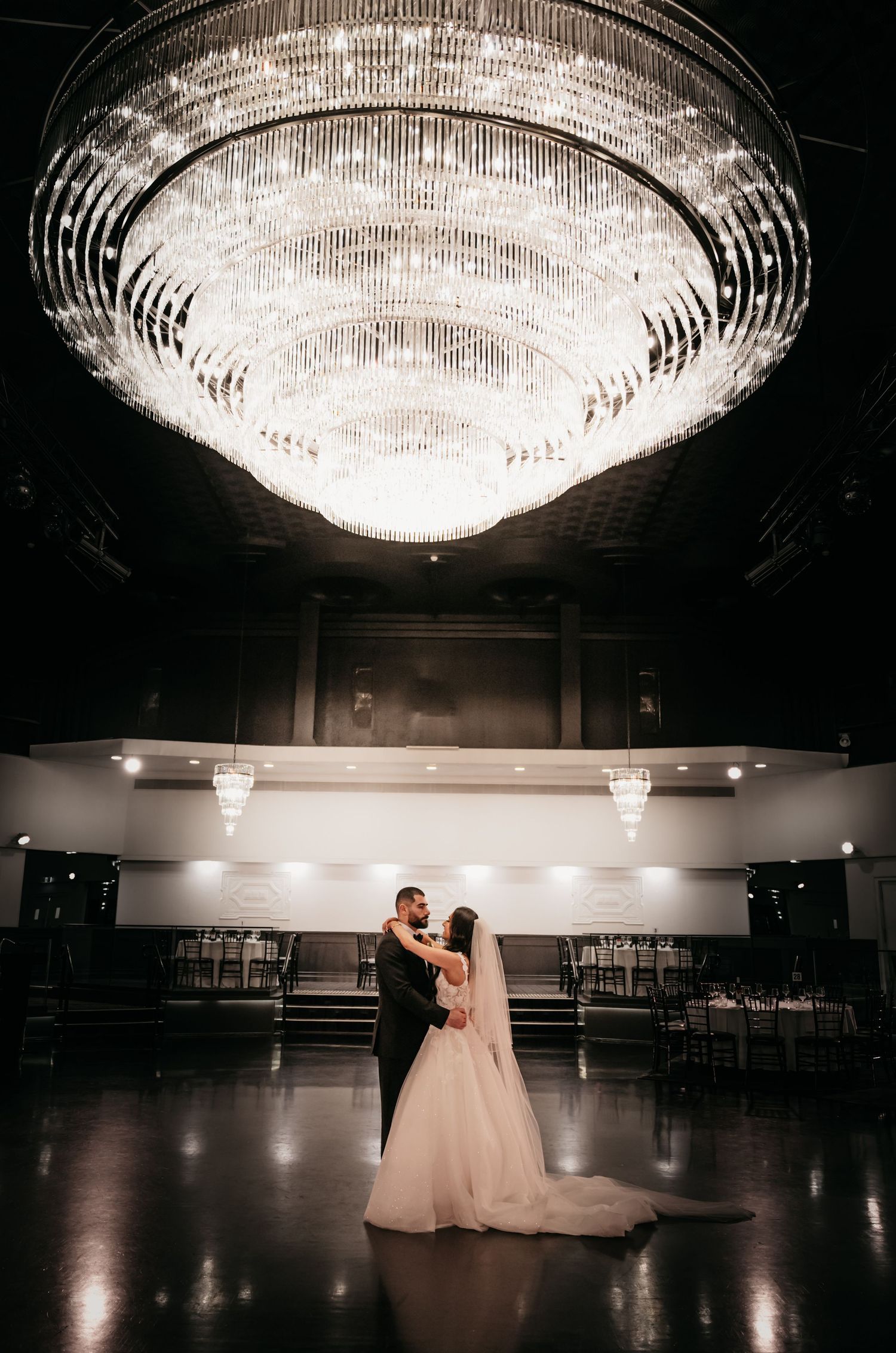 A grand crystal chandelier illuminates a spacious ballroom where two figures dance in elegant formal attire.