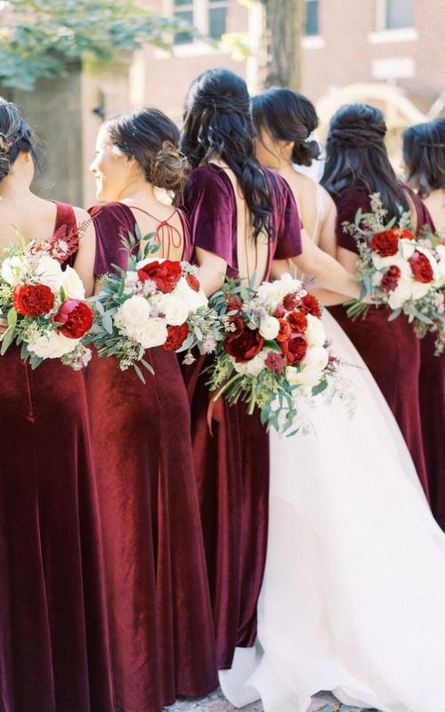 Bridesmaids in burgundy dresses hold red and white bouquets while standing in line with bride in white wedding gown.