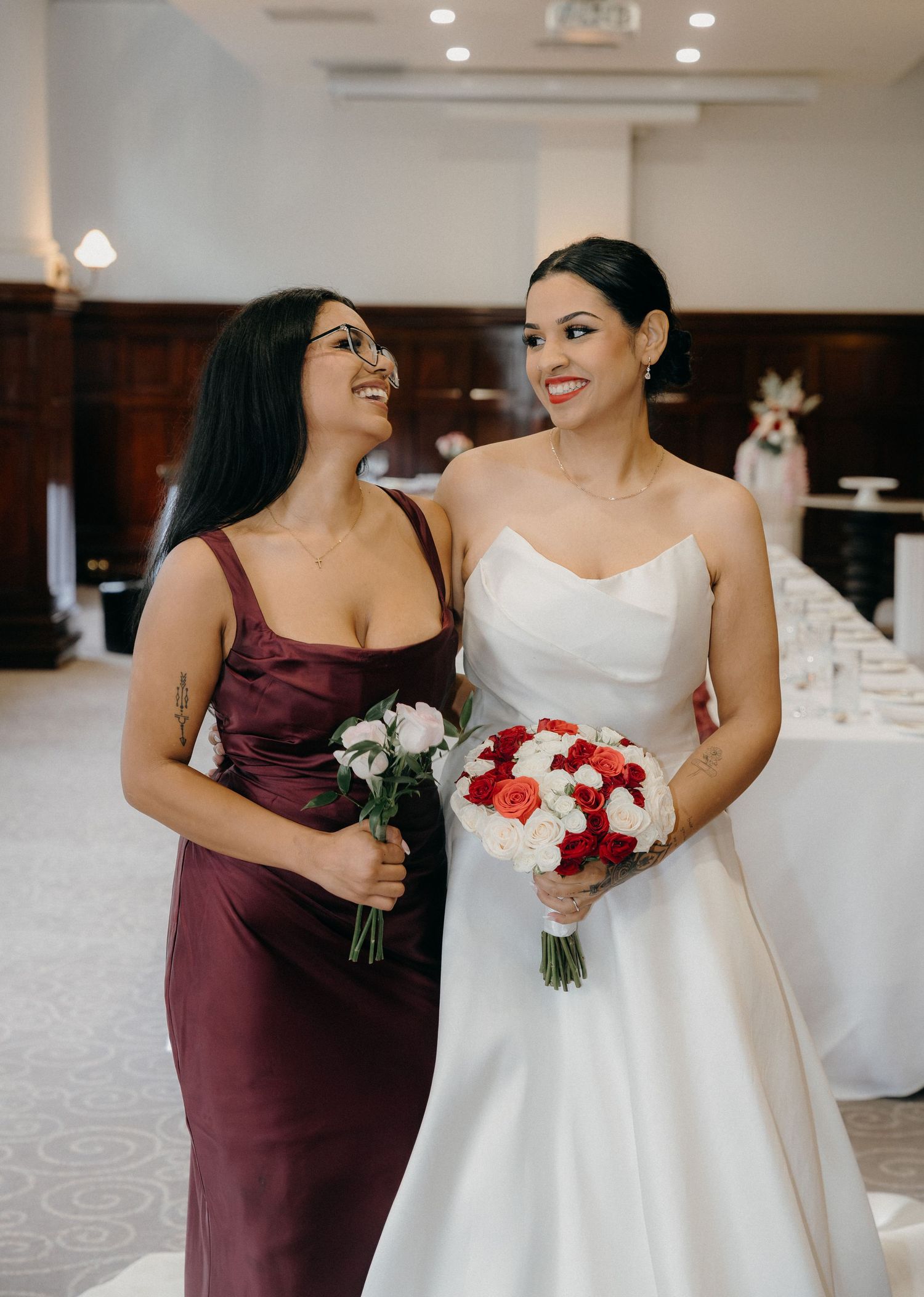 Bride in white strapless gown and bridesmaid in burgundy dress smiling, holding red and white bouquets at wedding reception.