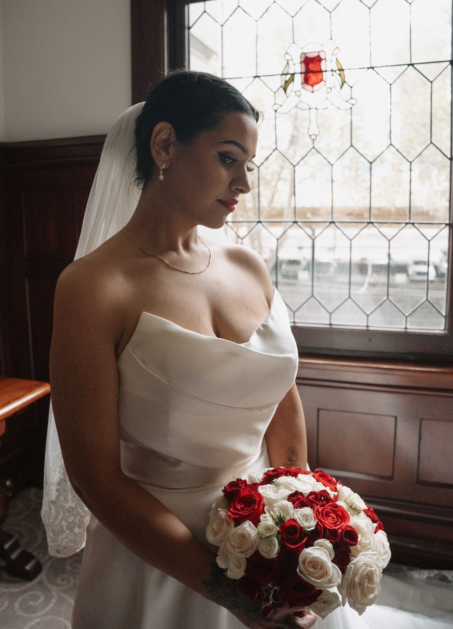 Bride in strapless white gown holding red and white rose bouquet, wearing veil, standing by stained glass window.