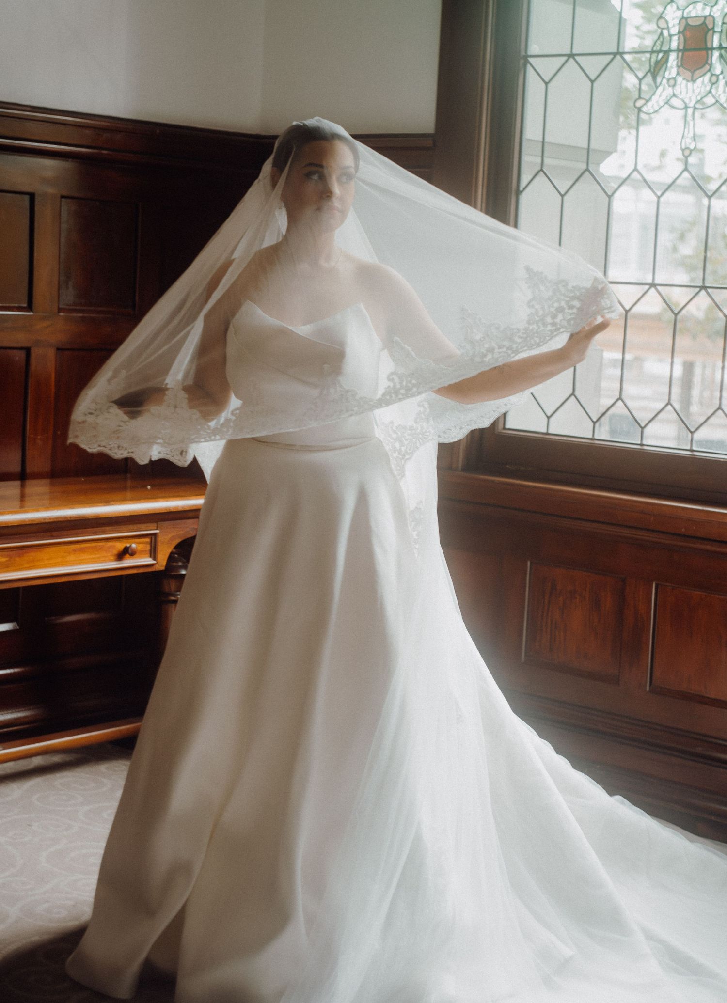 Bride in strapless white wedding gown with lace-trimmed cathedral veil posing near stained glass window.