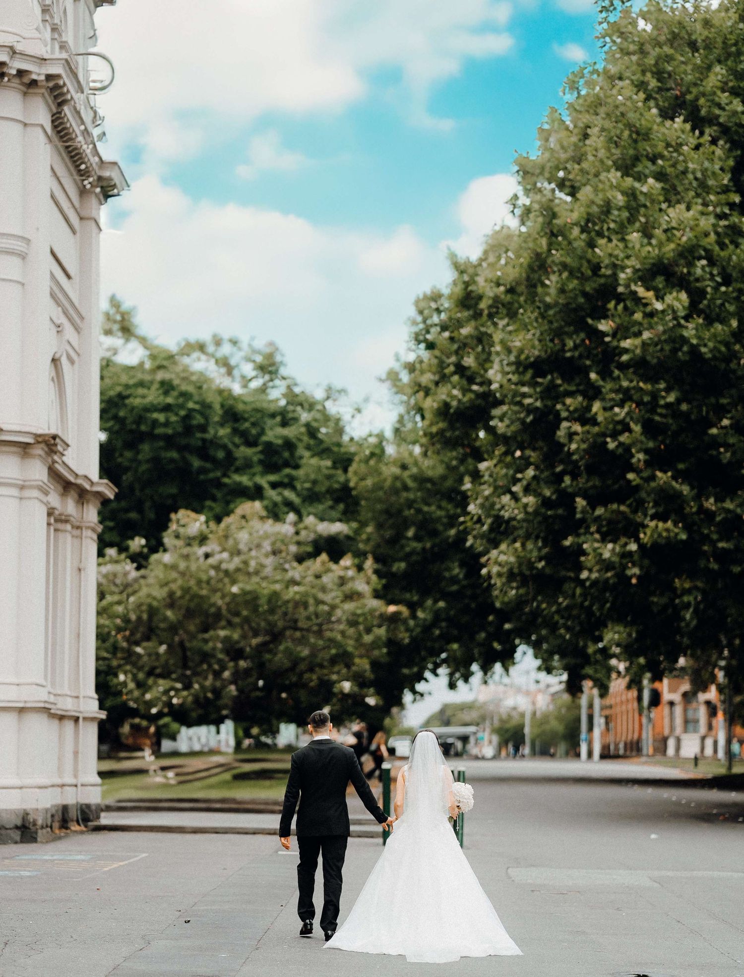 A couple in wedding attire walking away from camera on a tree-lined path near a white stone building.