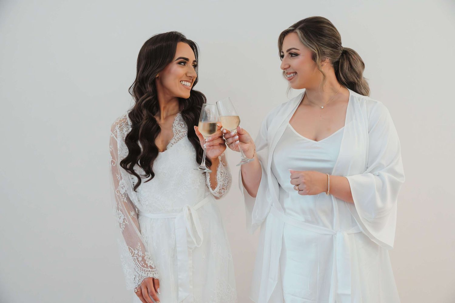 Two people in white lace robes share a cheerful moment together while getting ready.