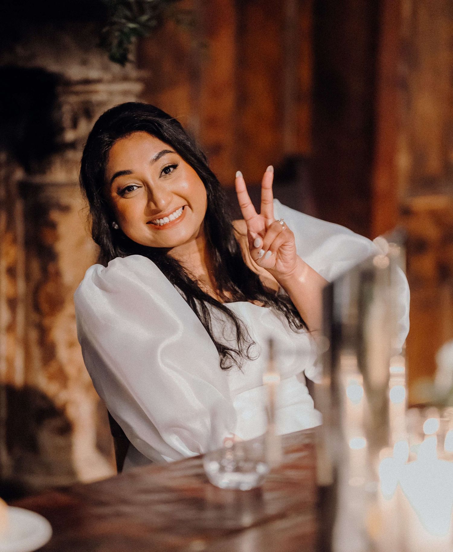 A person in a white top making a peace sign gesture while sitting at a table with a warm wooden backdrop.