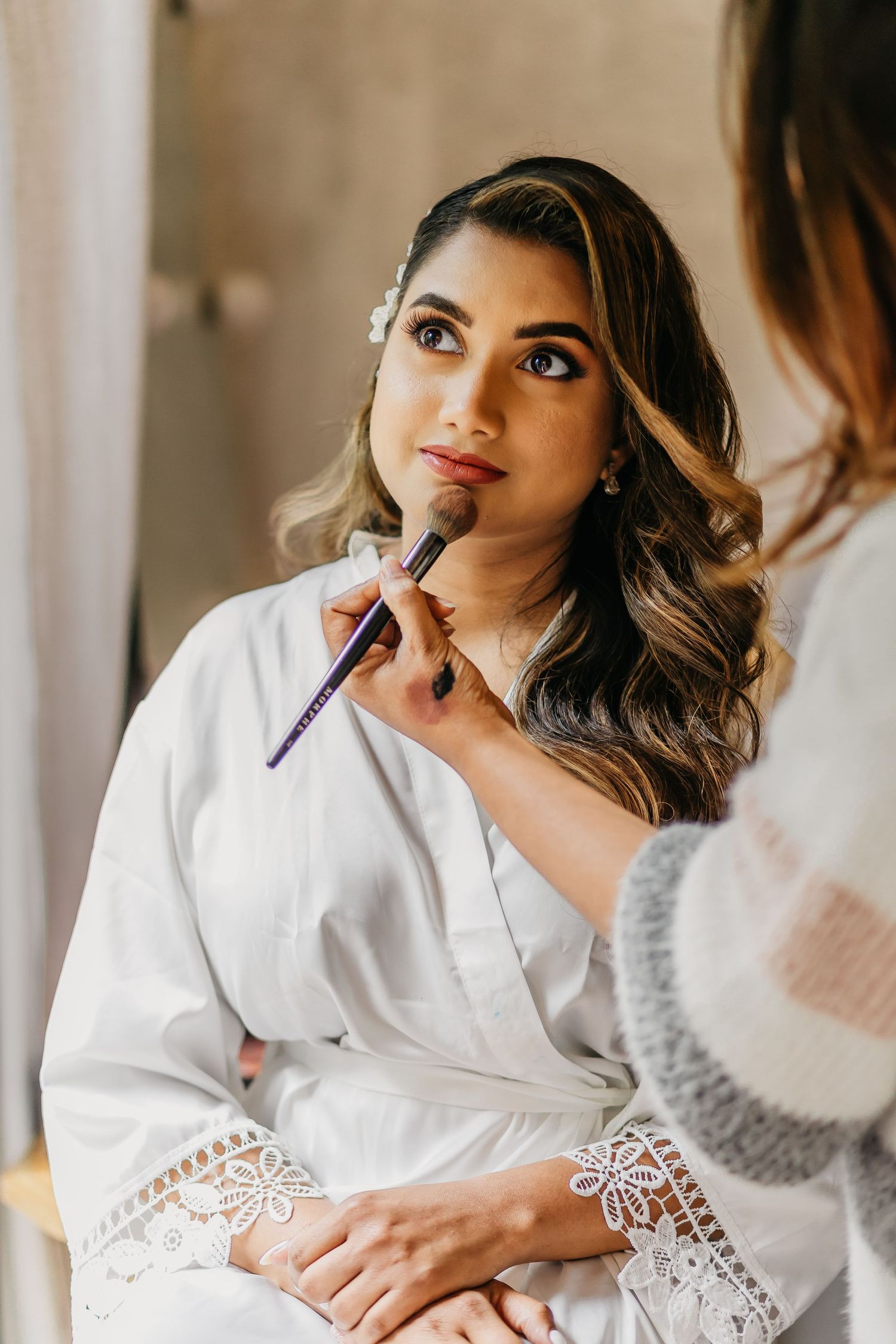 Professional makeup artist applying lipstick to client wearing white lace robe during bridal preparation.