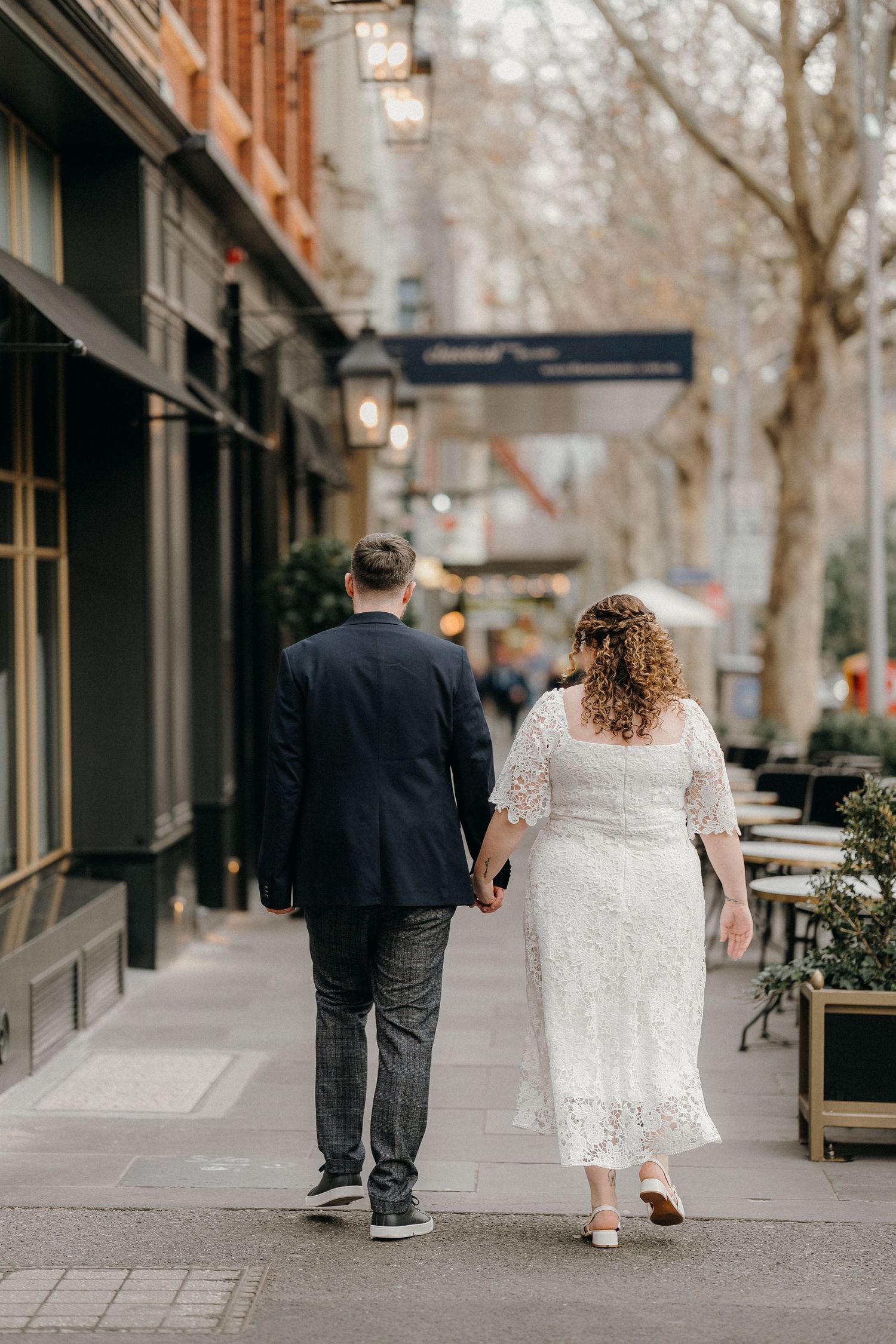 A couple in formal attire walks down a city sidewalk with storefronts and trees in winter.