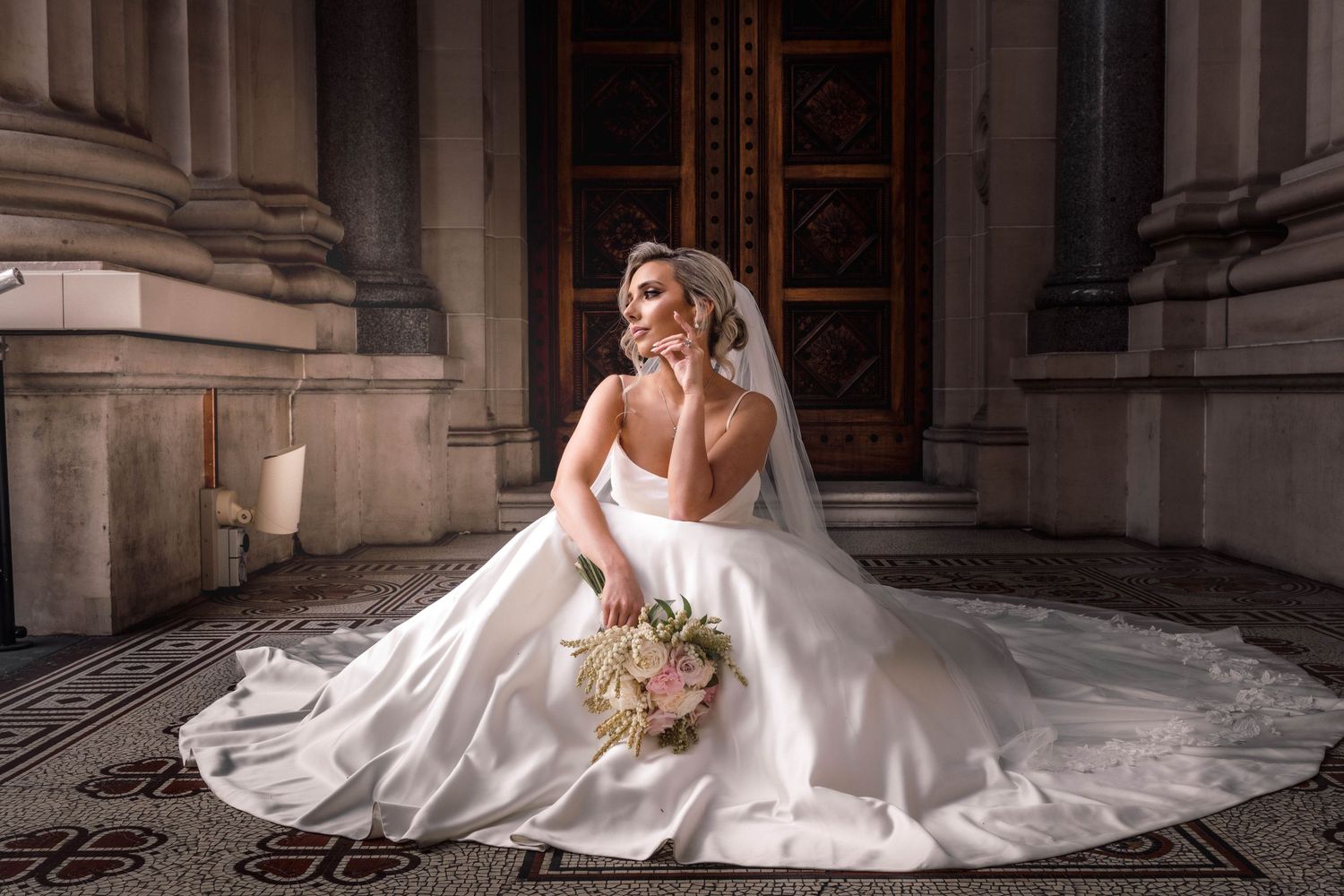 A bride in an elegant white satin gown with cathedral train sits gracefully on stone steps holding a neutral bouquet.