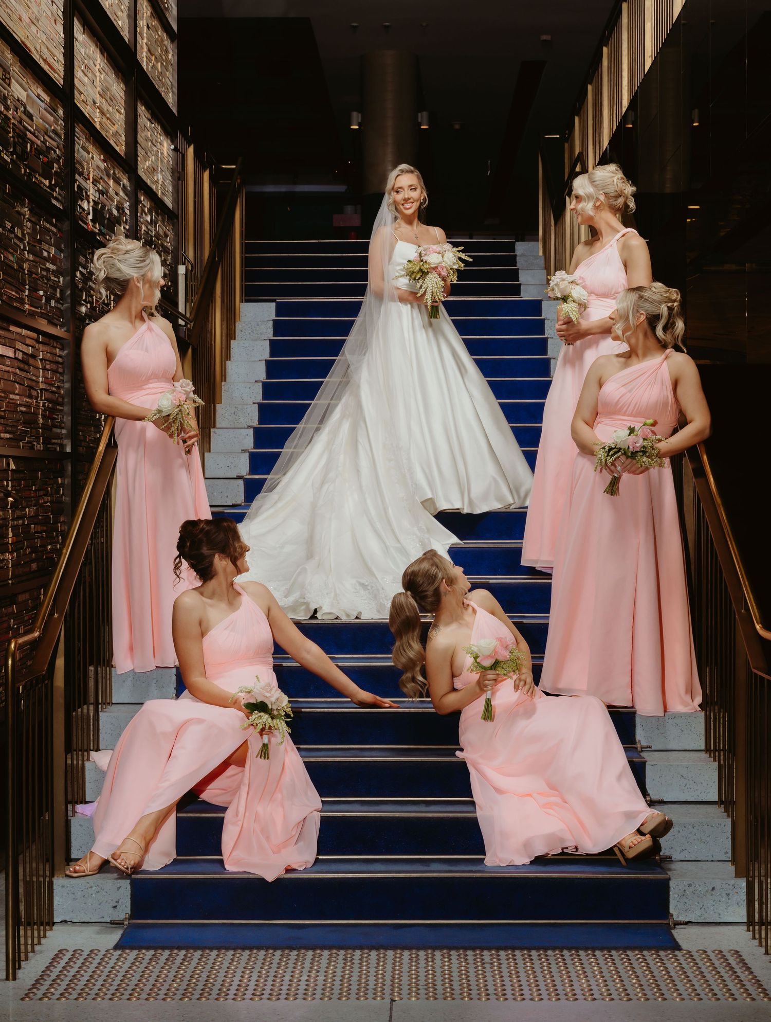 Bridesmaids in pink dresses pose playfully on blue carpeted stairs with bride in white gown during wedding photoshoot.