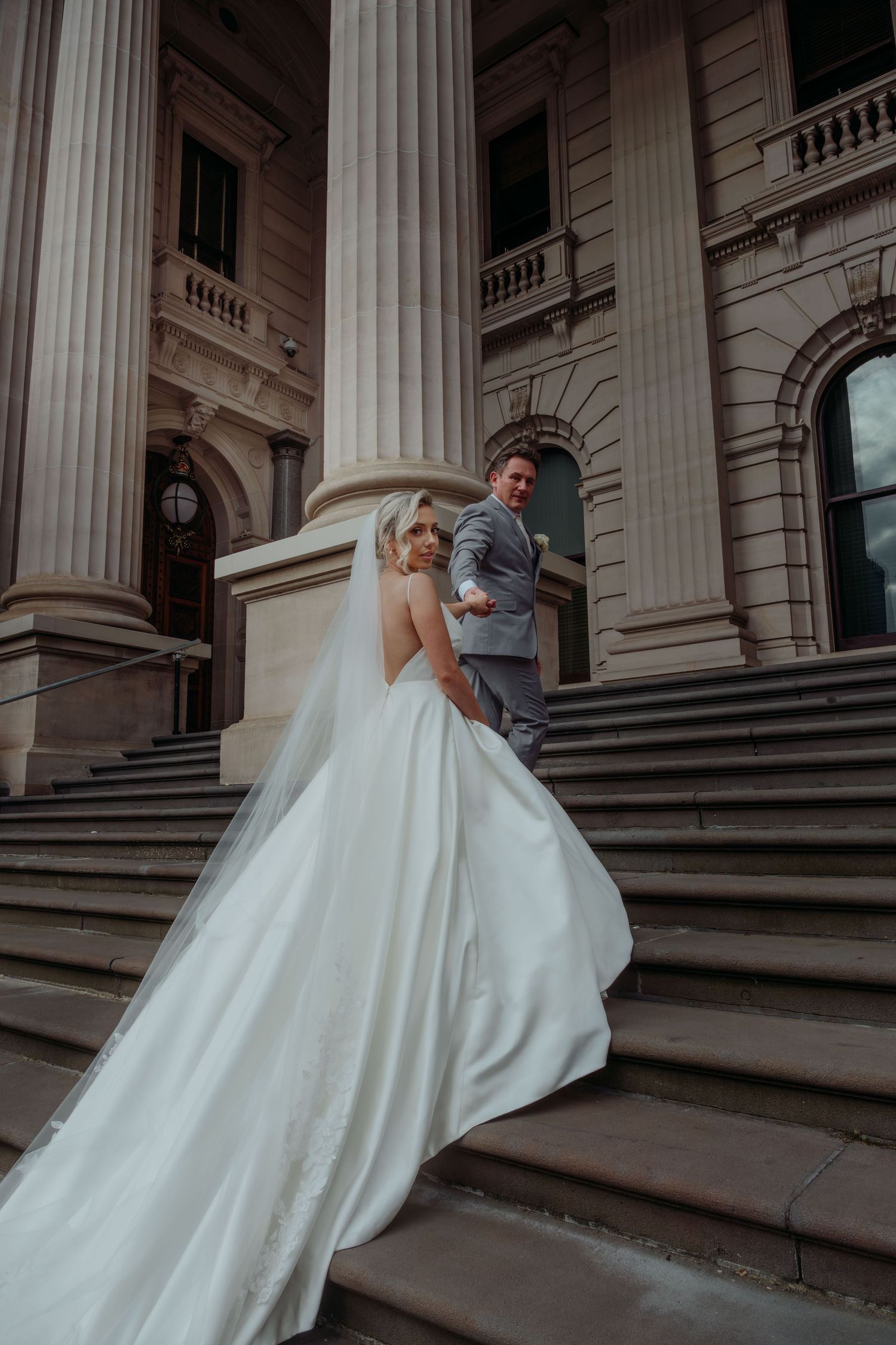 A couple in wedding attire pose on grand stone steps with towering classical columns of a historic building.