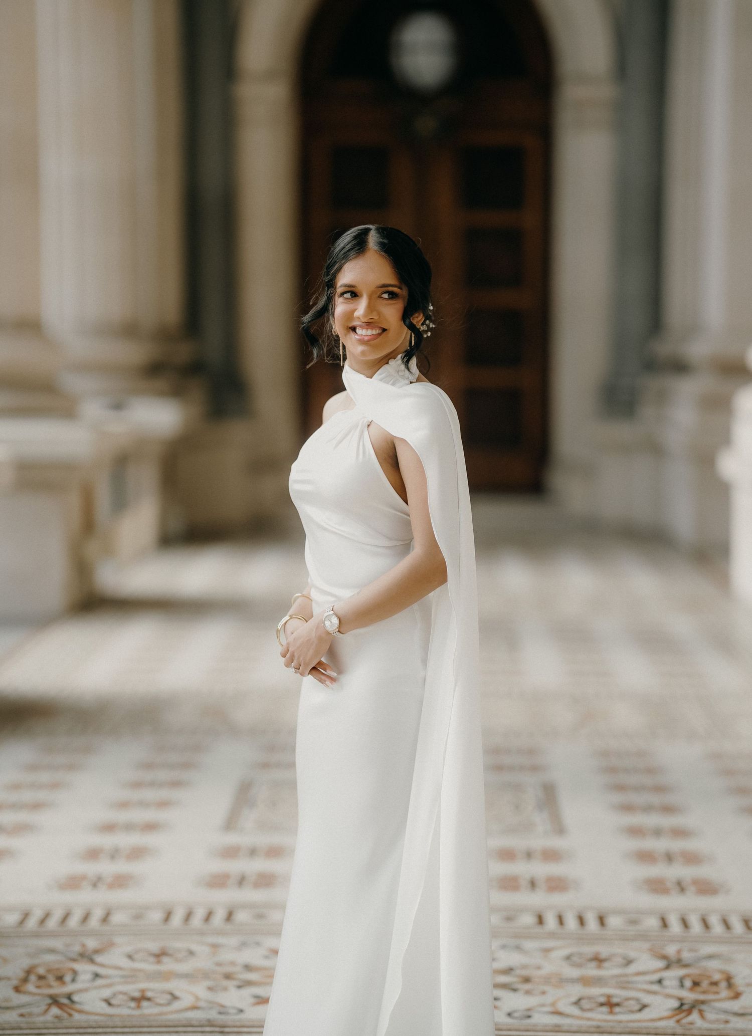 A bride in an elegant white dress smiles and twirls in a grand hallway with ornate tiled floors and columns.