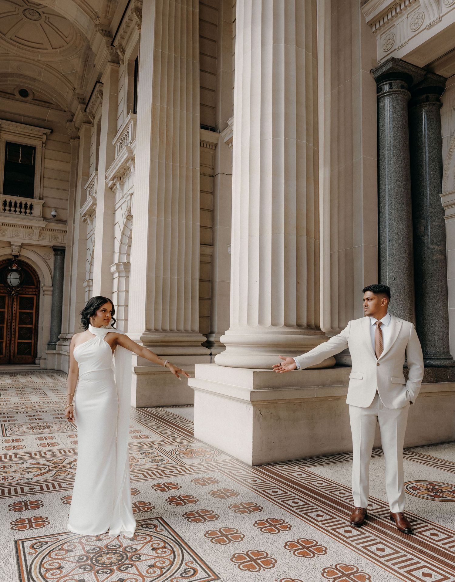 Elegant couple in white attire poses gracefully in a grand marble hallway with ornate tiled floors and classical columns.