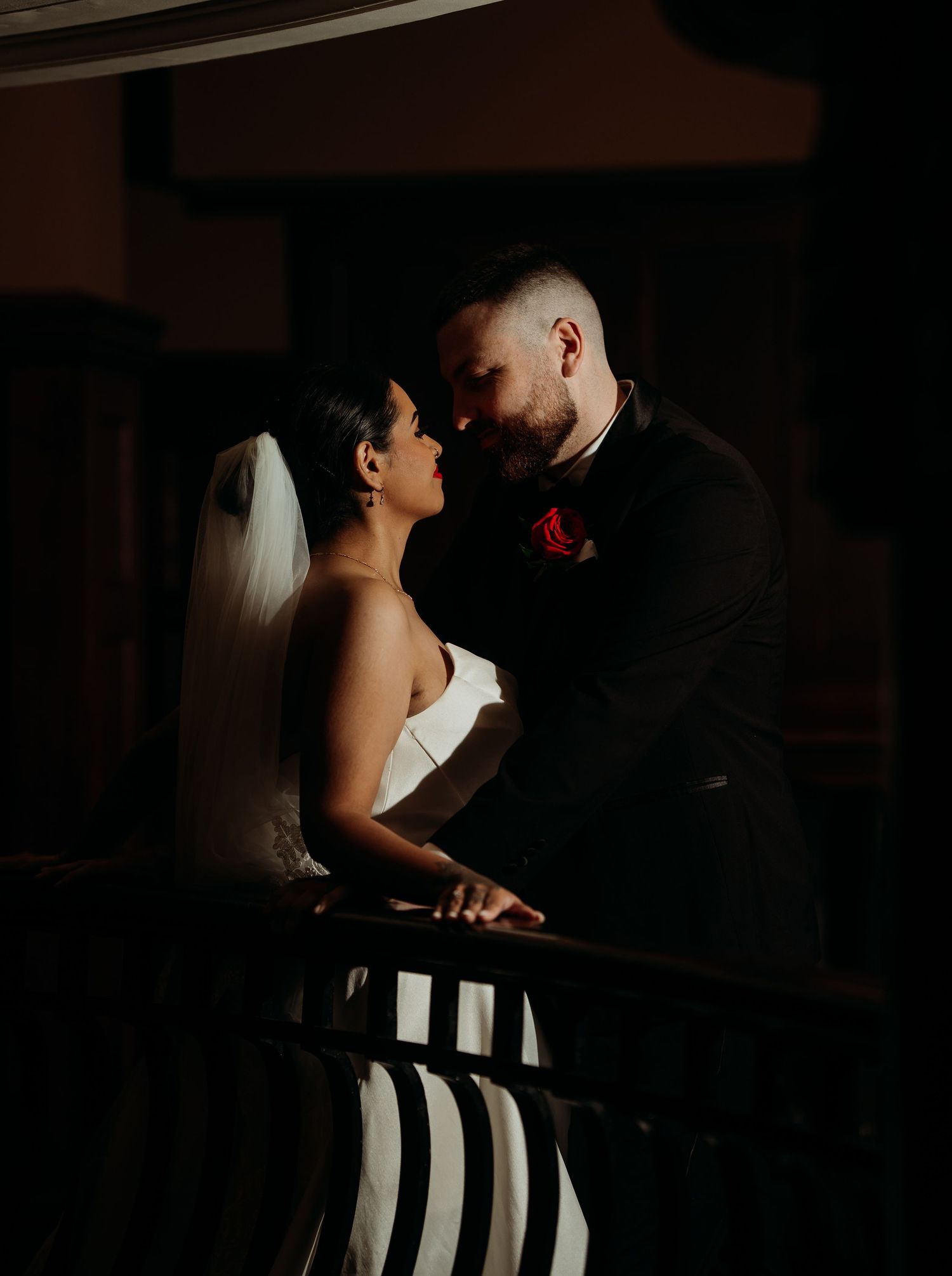 A bride and groom share a romantic moment on a dimly lit balcony, the groom wearing a red rose boutonniere.
