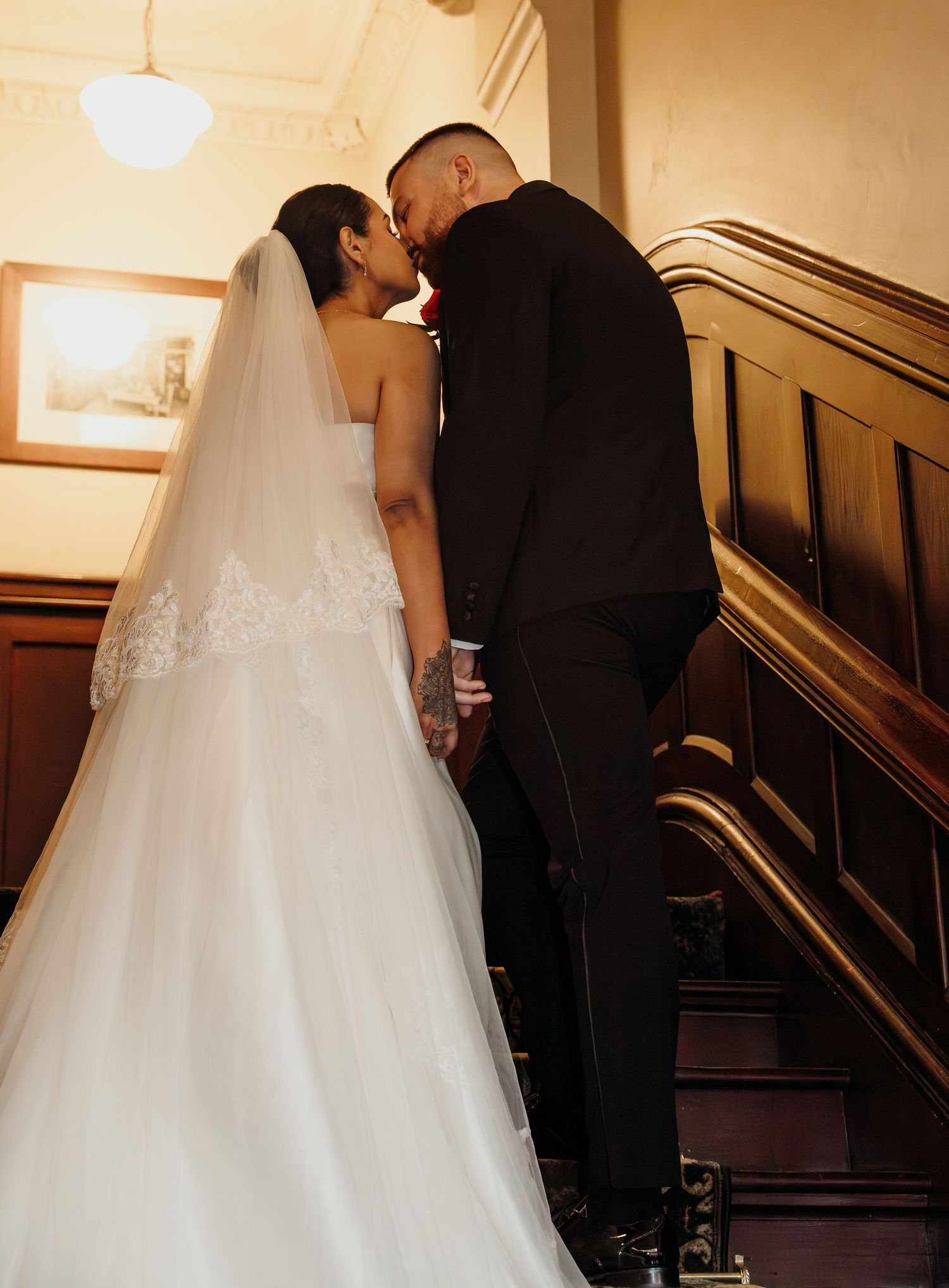 Bride in white gown and veil sharing a romantic kiss with groom in black suit on wooden staircase.