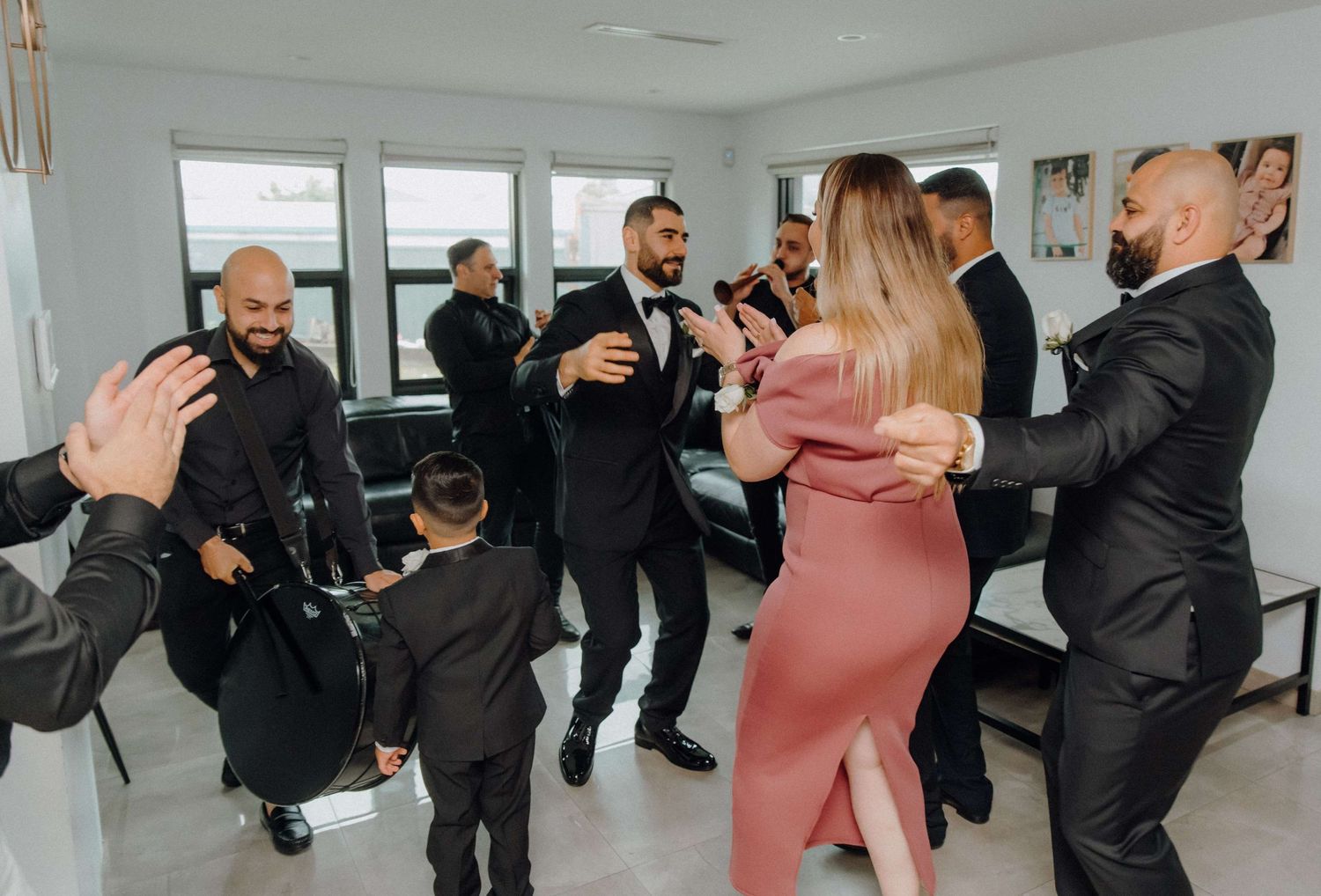 A group dressed in formal attire socialize and dance together in a bright room with windows.