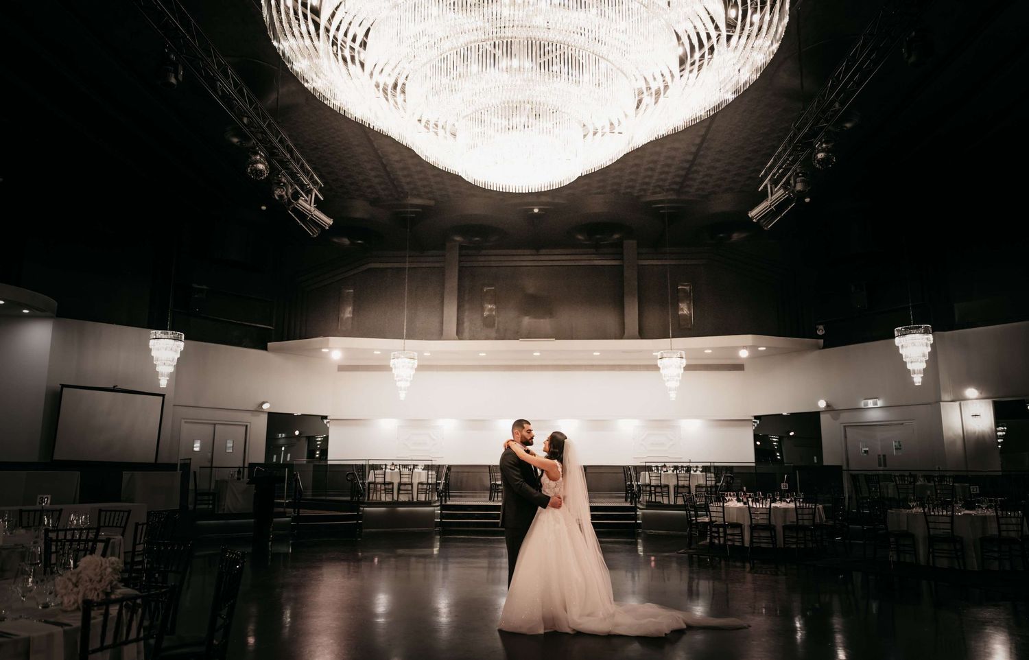 A couple shares their first dance under a glowing chandelier in an elegant ballroom reception venue.
