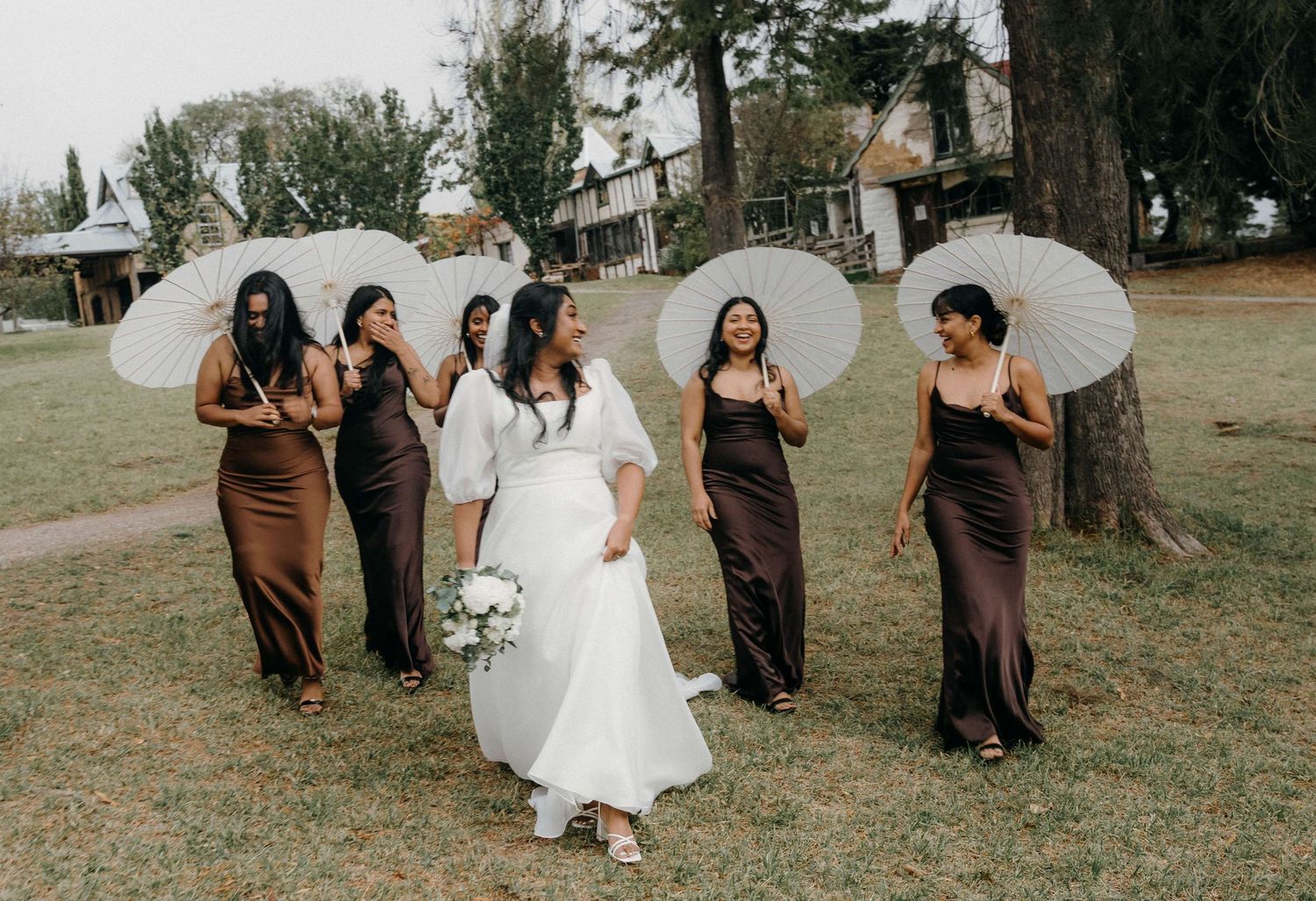 Wedding party walking outdoors on grass holding white parasol umbrellas in brown and white formal dresses.