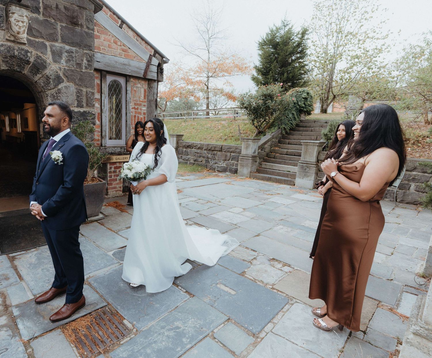 Outdoor wedding scene with couple in formal attire standing on stone patio beside historic brick building.