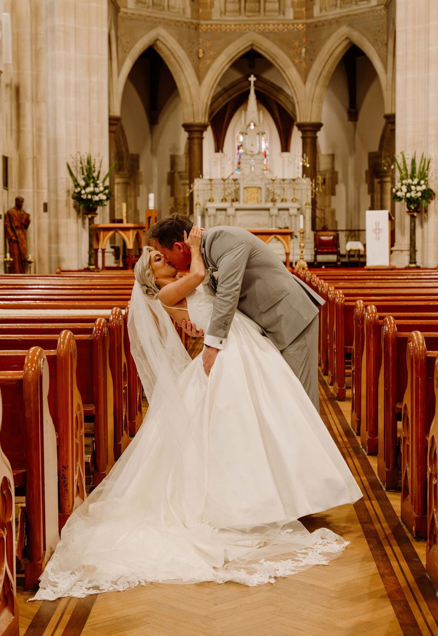 A bride and groom share a romantic kiss in a grand cathedral church aisle with wooden pews and Gothic architecture.