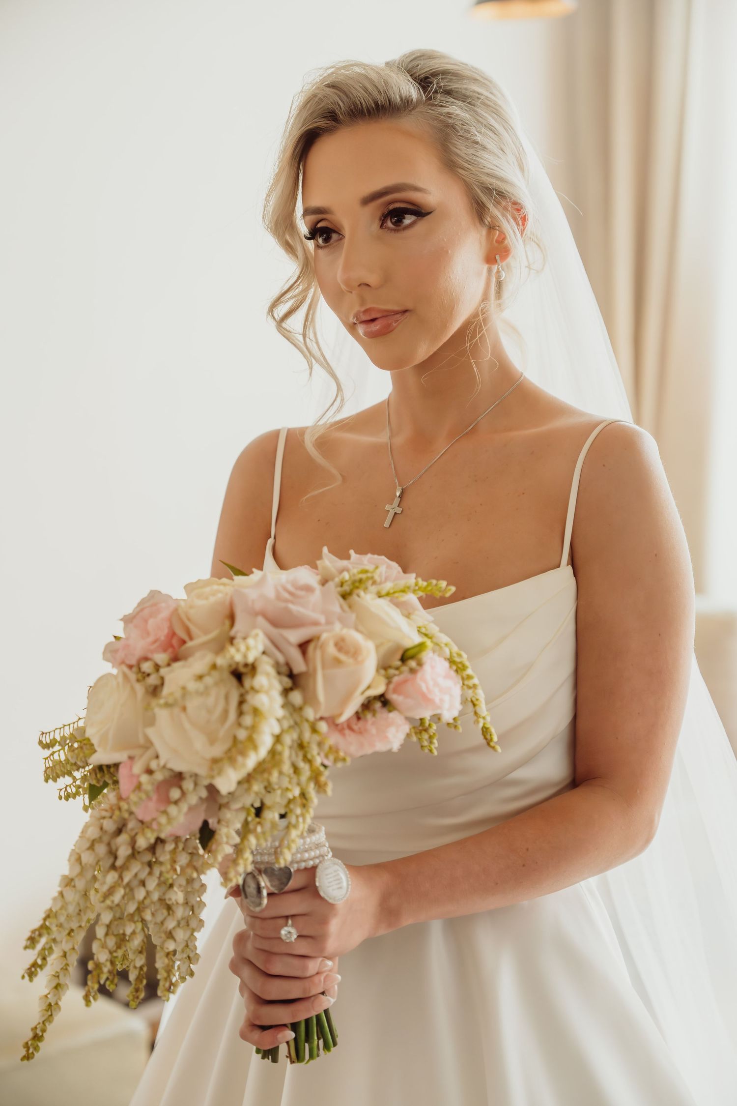 A bride in a white dress holds a romantic bouquet of blush pink roses and delicate greenery against a light background.