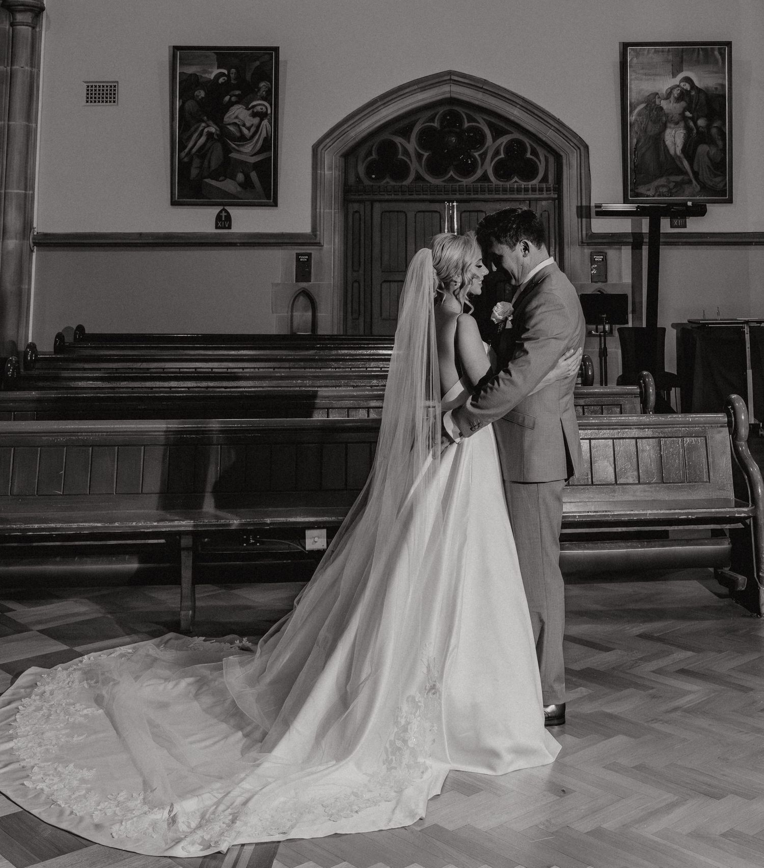 A romantic black and white wedding photograph showing a couple embracing in a church with wooden pews and religious artwork.