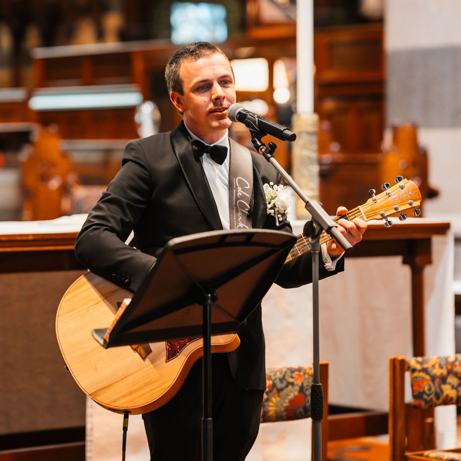 A musician in a black suit performs with an acoustic guitar in a warmly lit church or concert hall setting.