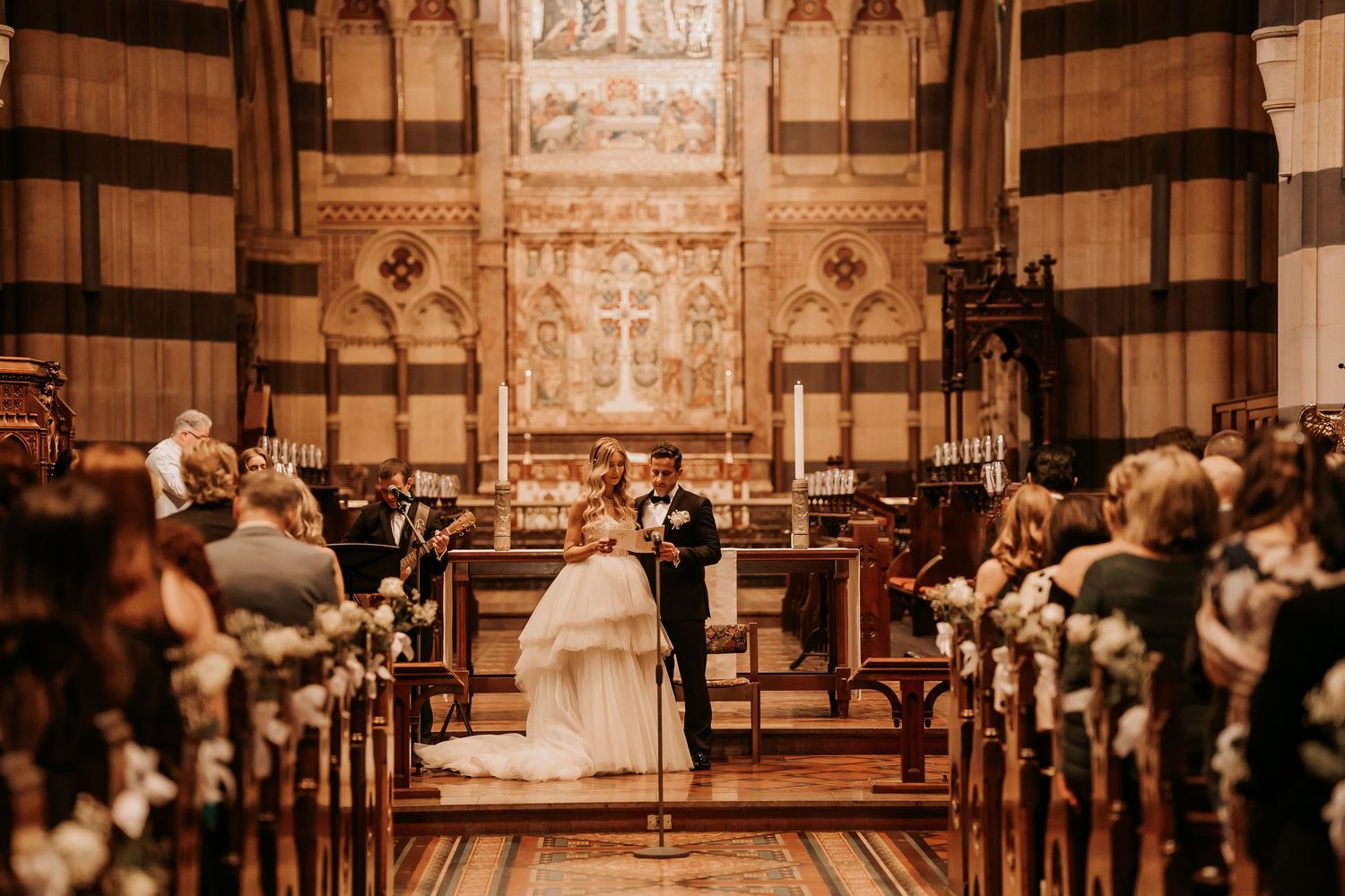 A warm-toned church wedding ceremony with guests seated in wooden pews and decorative floral arrangements along the aisle.