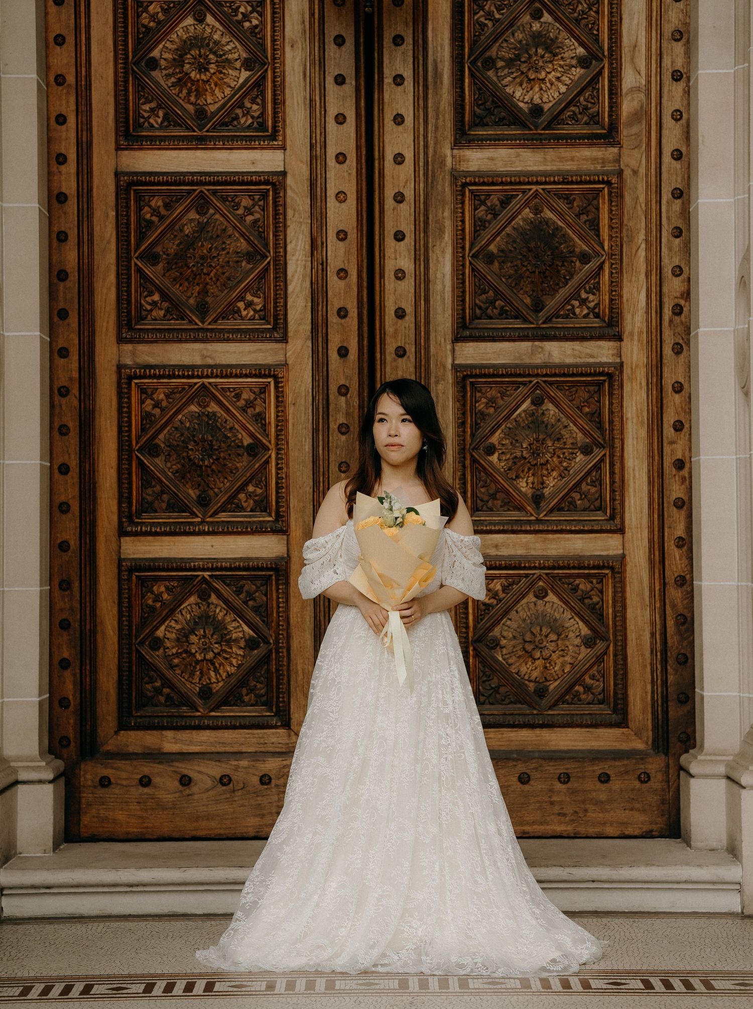 Elegant white lace wedding dress photographed against ornate wooden doors with geometric patterns.