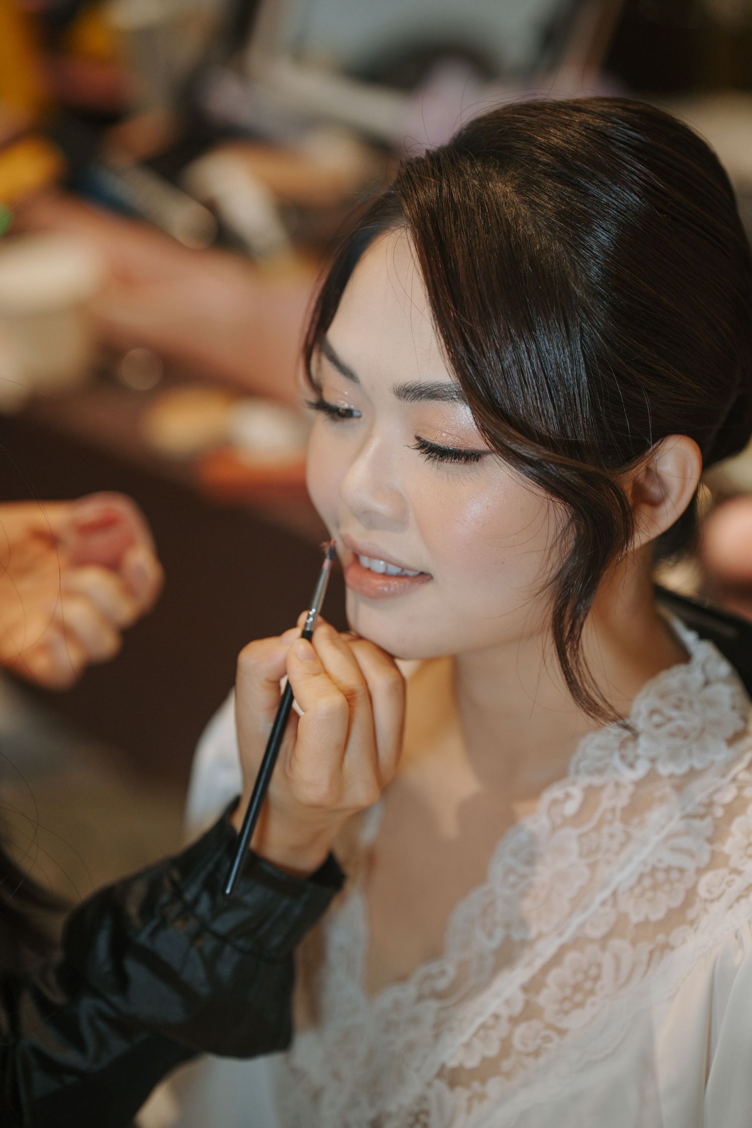 A makeup artist applies cosmetics to a bride wearing a white lace dress during wedding preparations.
