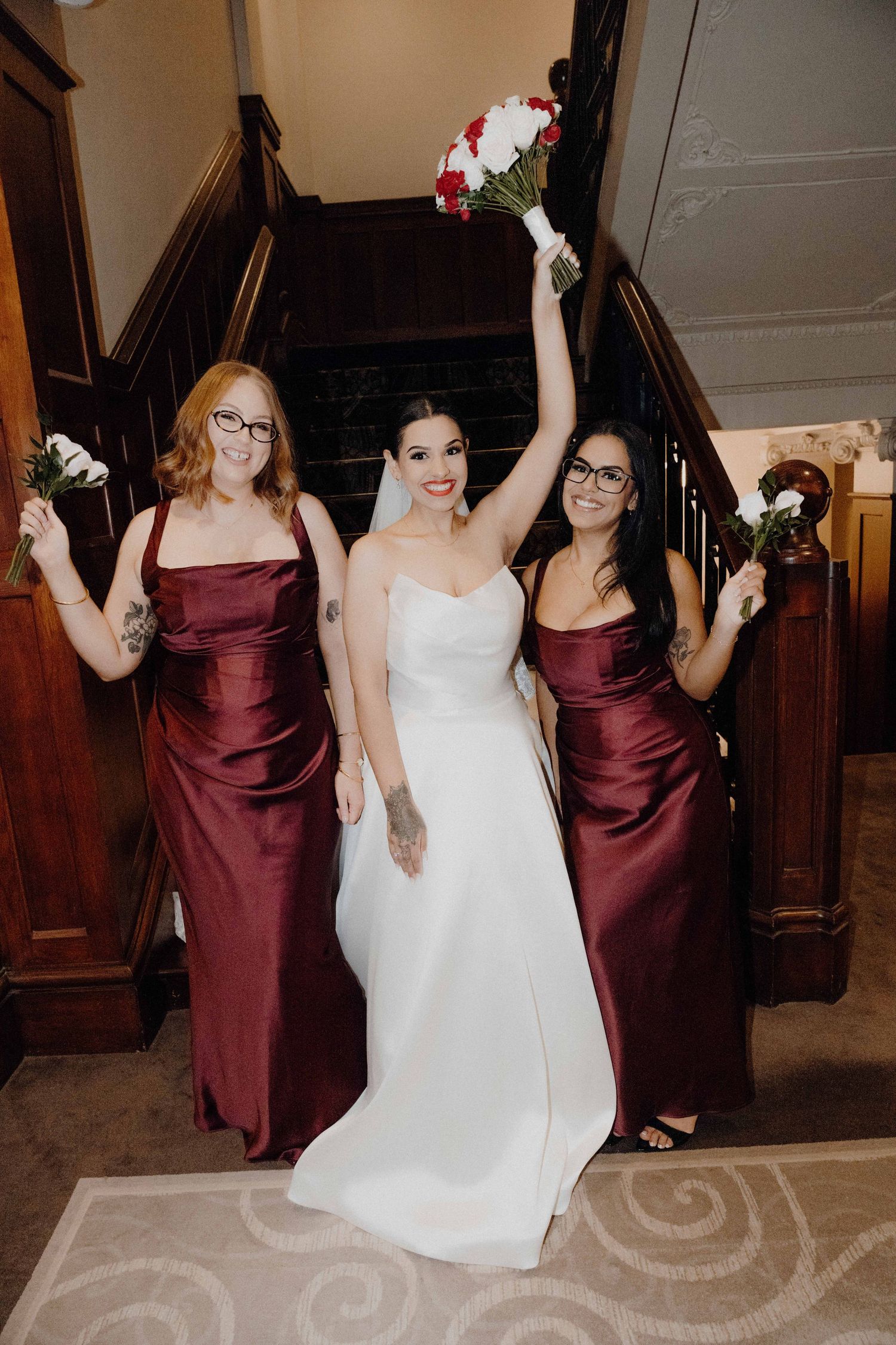 Bride in white gown poses with two bridesmaids in burgundy satin dresses, holding bouquets on a staircase.