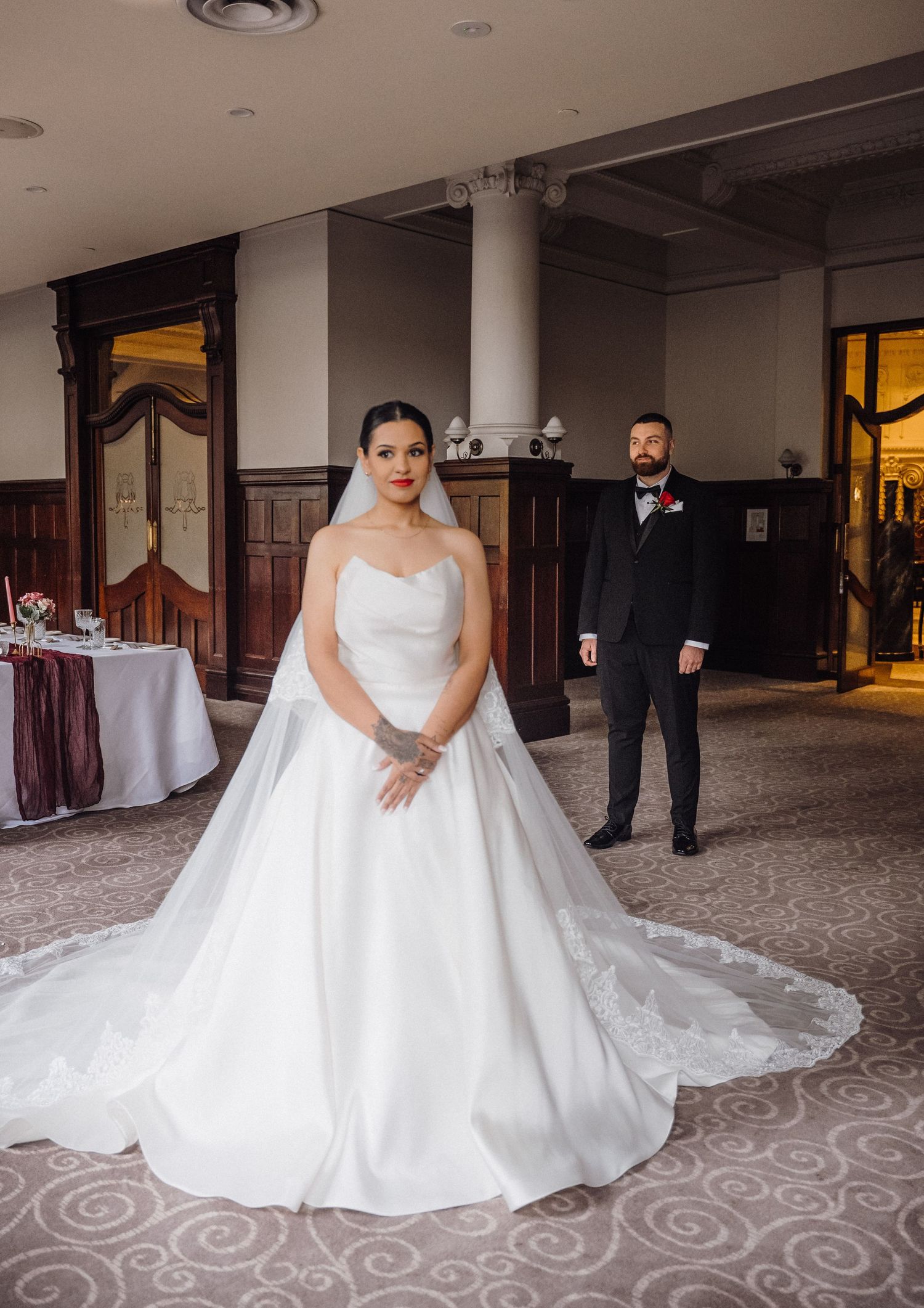 Bride in elegant white ball gown with long train stands before groom in black tuxedo in ornate wedding venue.