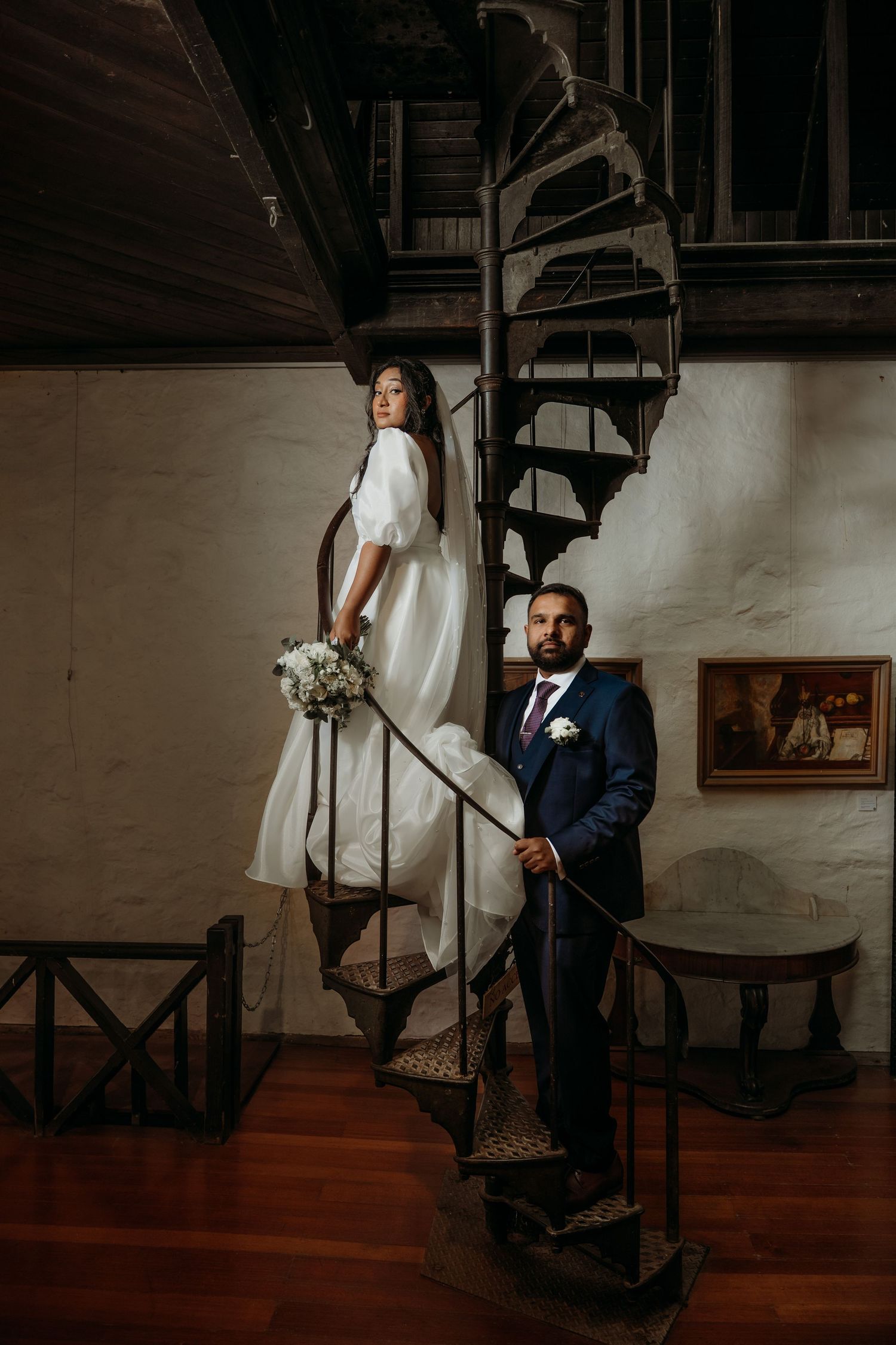 A romantic wedding photo on a spiral staircase features a white dress and navy suit in a rustic indoor setting.