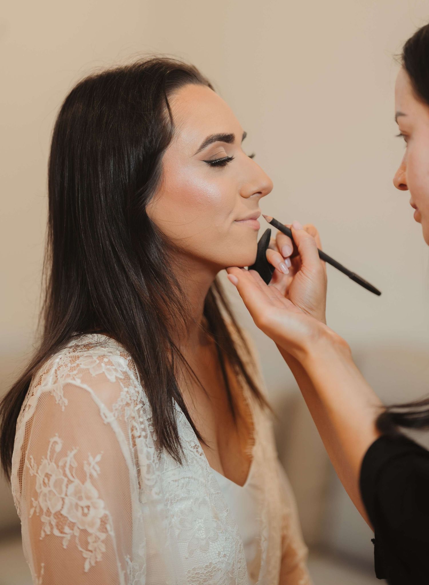 A makeup artist applies cosmetics to a bride wearing a delicate white lace gown during wedding preparations.