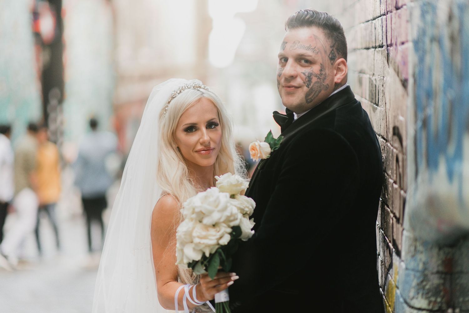 Newlyweds share a tender moment against a brick wall while holding a bouquet of white flowers.