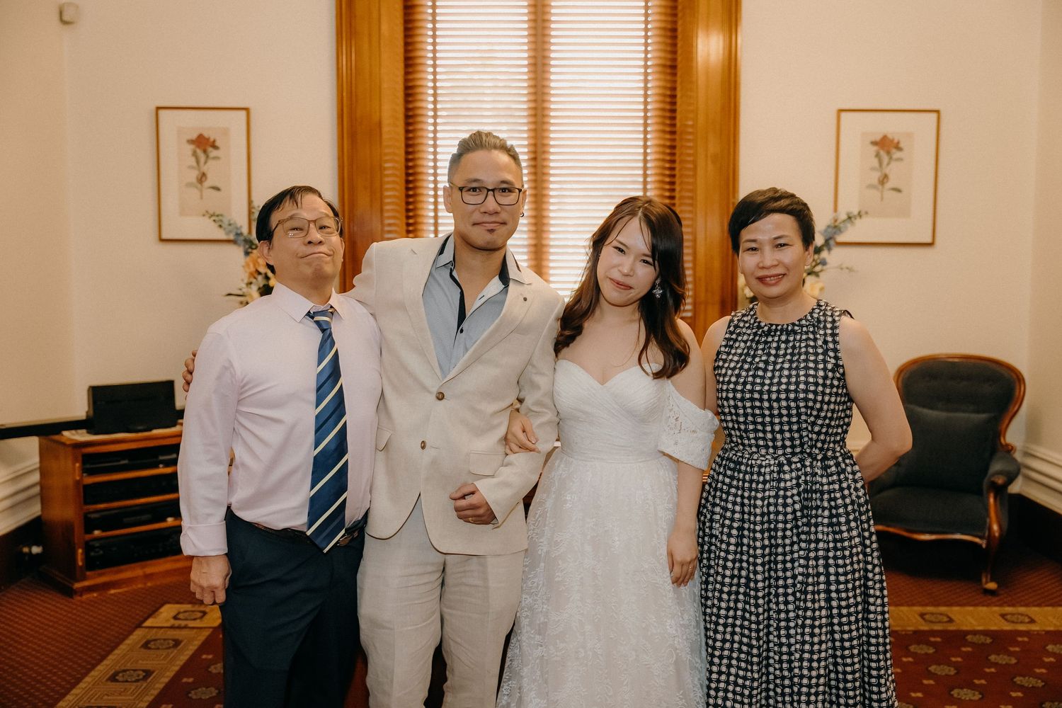 A group poses together in formal attire including a white wedding dress and light suit against a warm-toned interior.