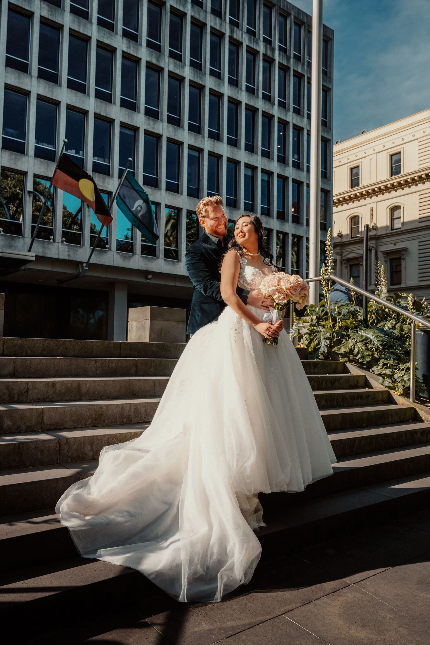 A newlywed couple embrace on outdoor stone steps with urban architecture and flags in the background.