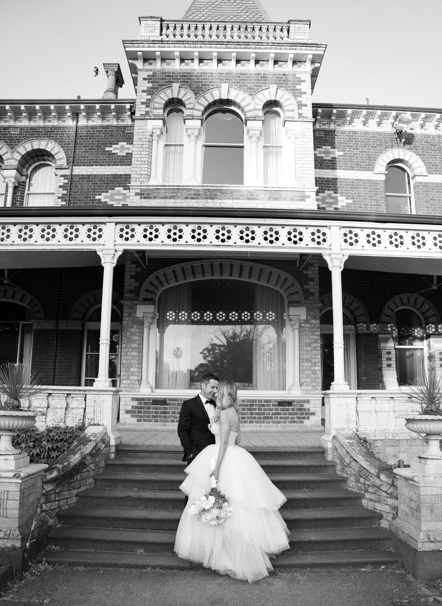 A couple stands on the steps of a grand Victorian-style mansion with ornate brick architecture and arched windows.