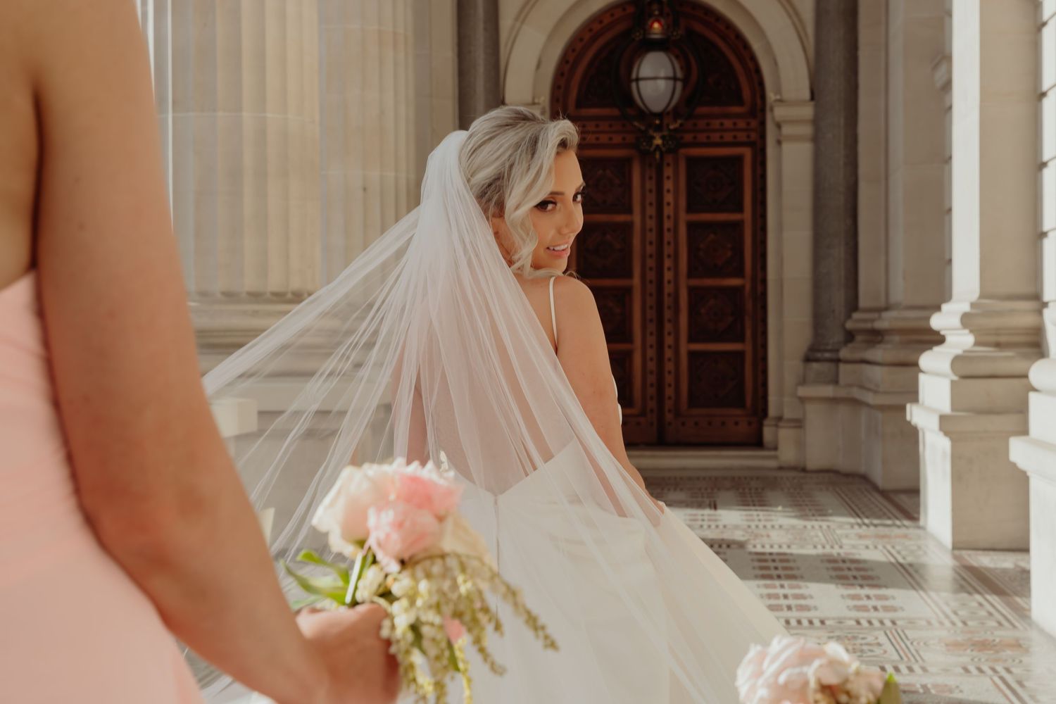 A bride in a flowing white veil stands near ornate wooden doors at an elegant venue with stone columns.