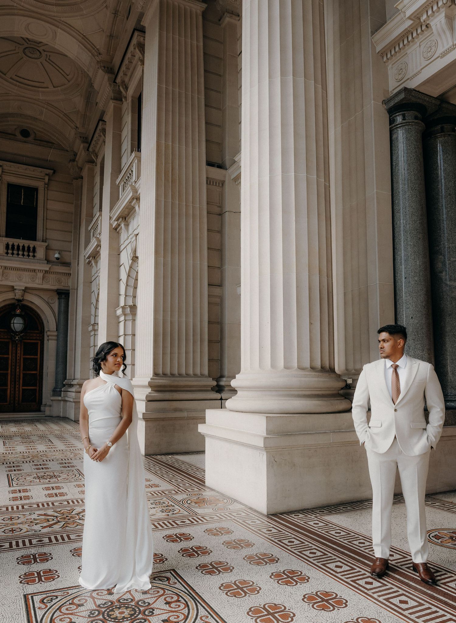 A couple in elegant white wedding attire pose between classical stone columns with ornate floor patterns.