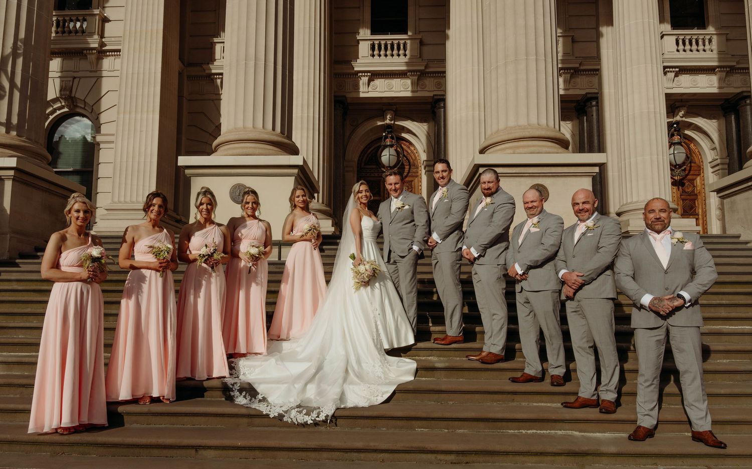 Wedding party in pink bridesmaid dresses and gray suits poses on grand stone steps of classical building.