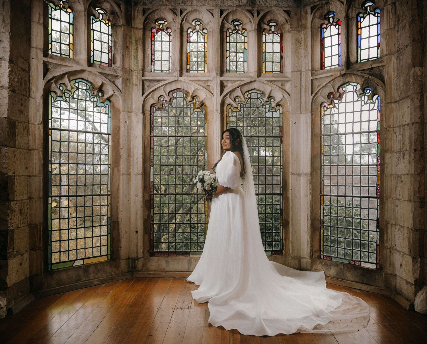 Elegant white wedding gown silhouetted in a Gothic stone chapel with tall arched windows and natural light streaming through.