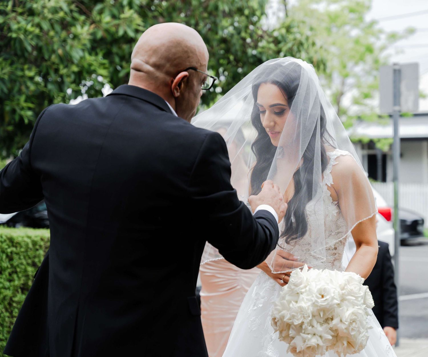 A tender moment captured during a wedding ceremony as a person adjusts the bride's delicate veil outdoors.