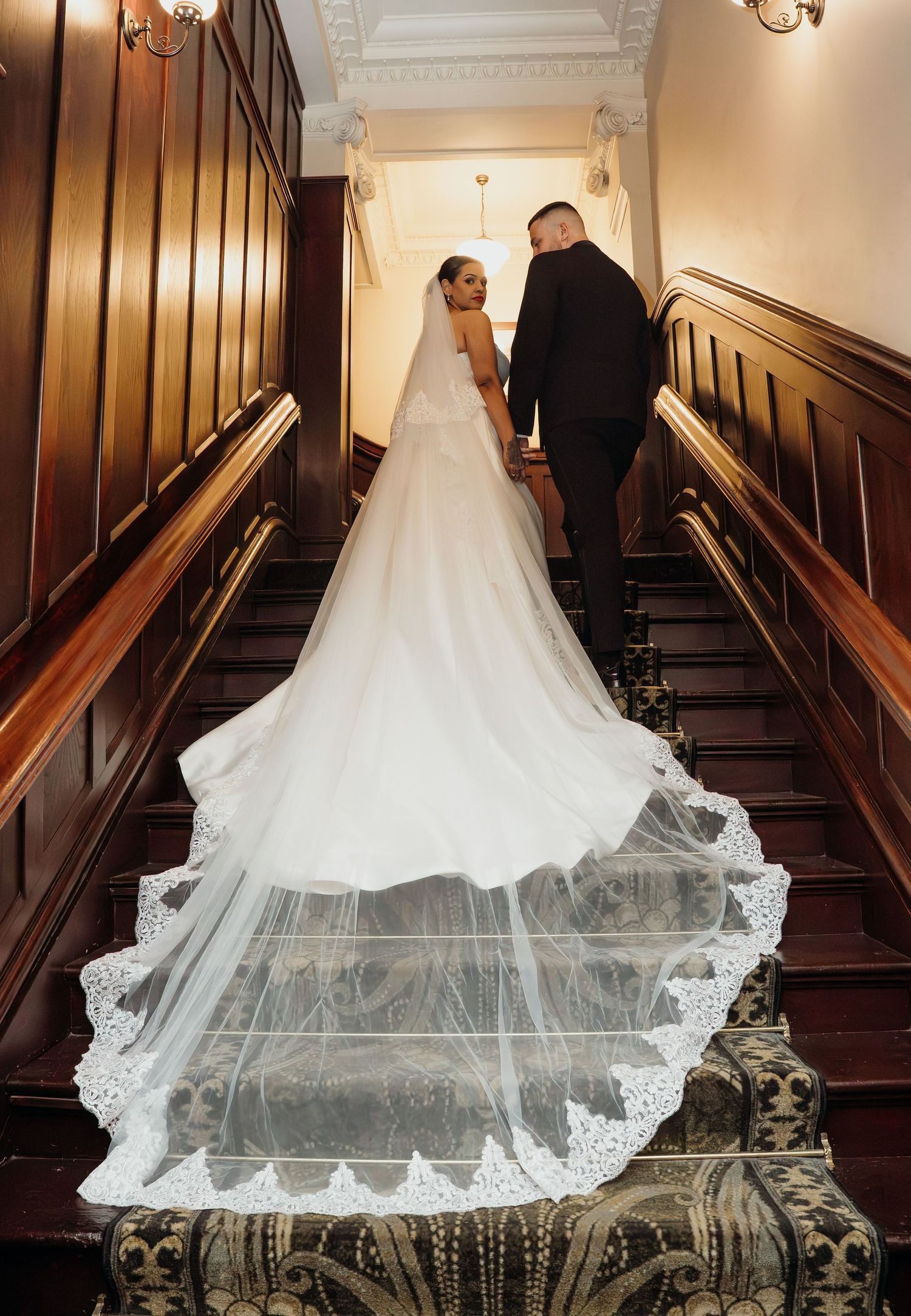 Bride in white gown with lace cathedral veil and groom in black suit ascending elegant wooden staircase.
