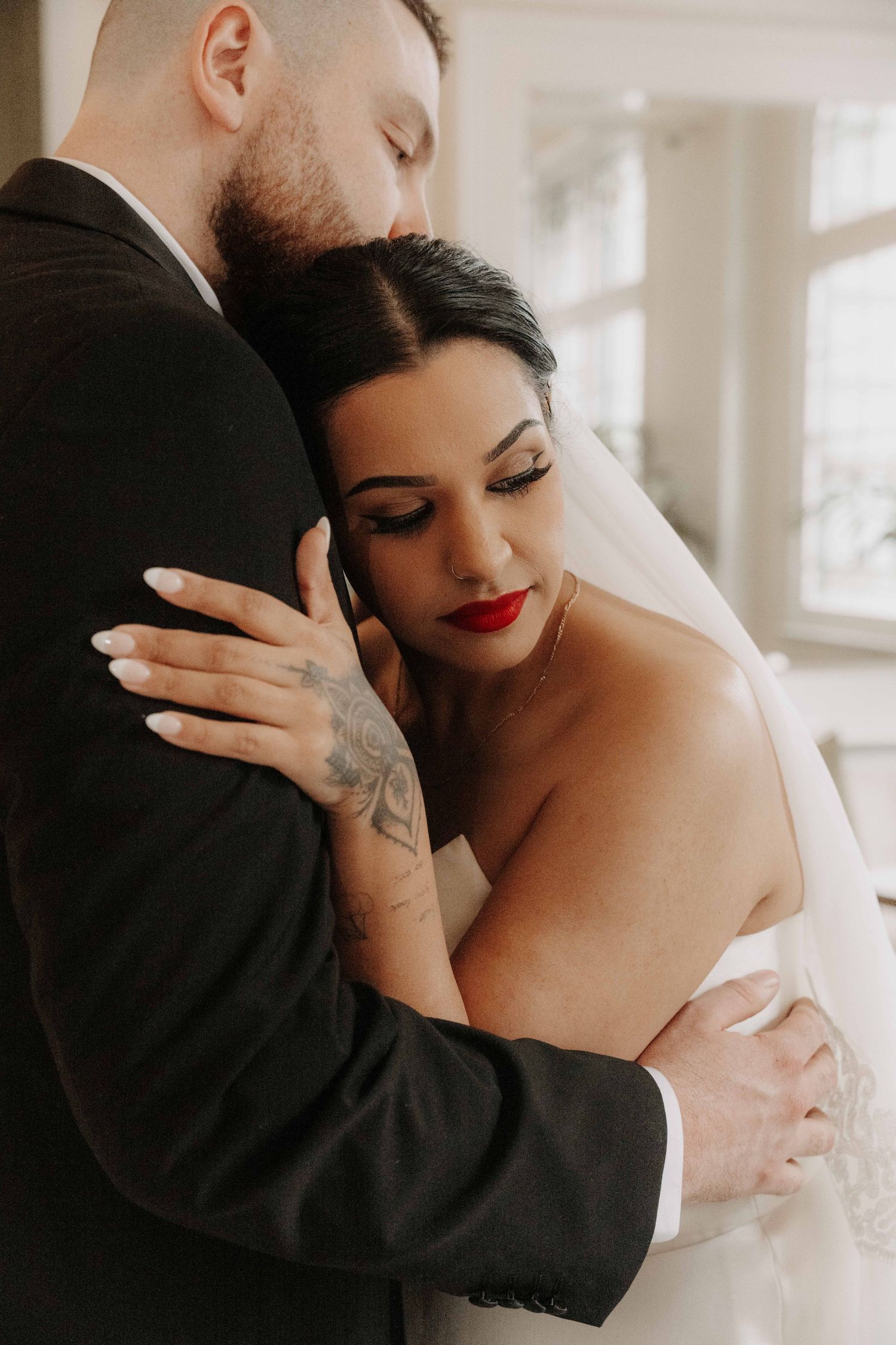 Bride with red lips and tattoos embraces groom in black suit during intimate wedding portrait.