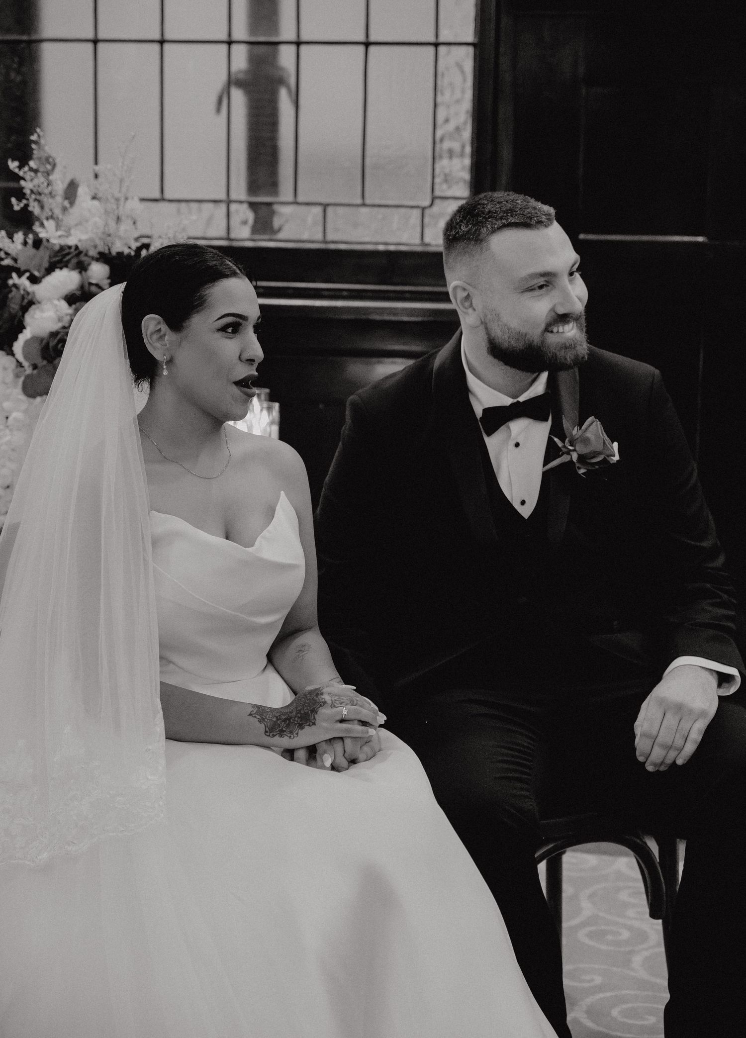 Black and white photo of a bride in white gown and groom in tuxedo seated together during their wedding ceremony.