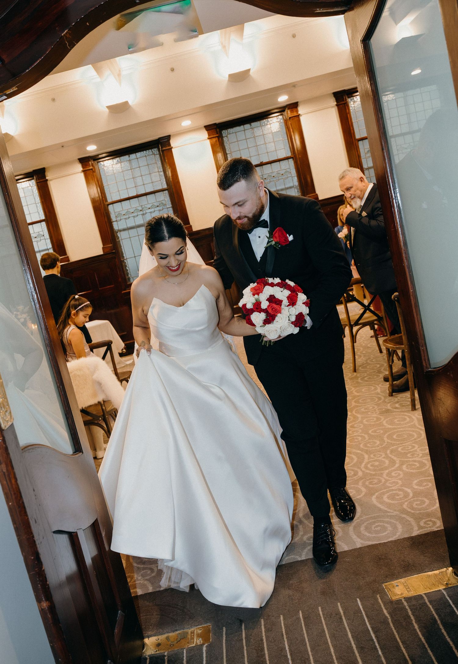 Bride and groom walk arm in arm through venue doorway, bride holding red and white bouquet in elegant white ballgown.