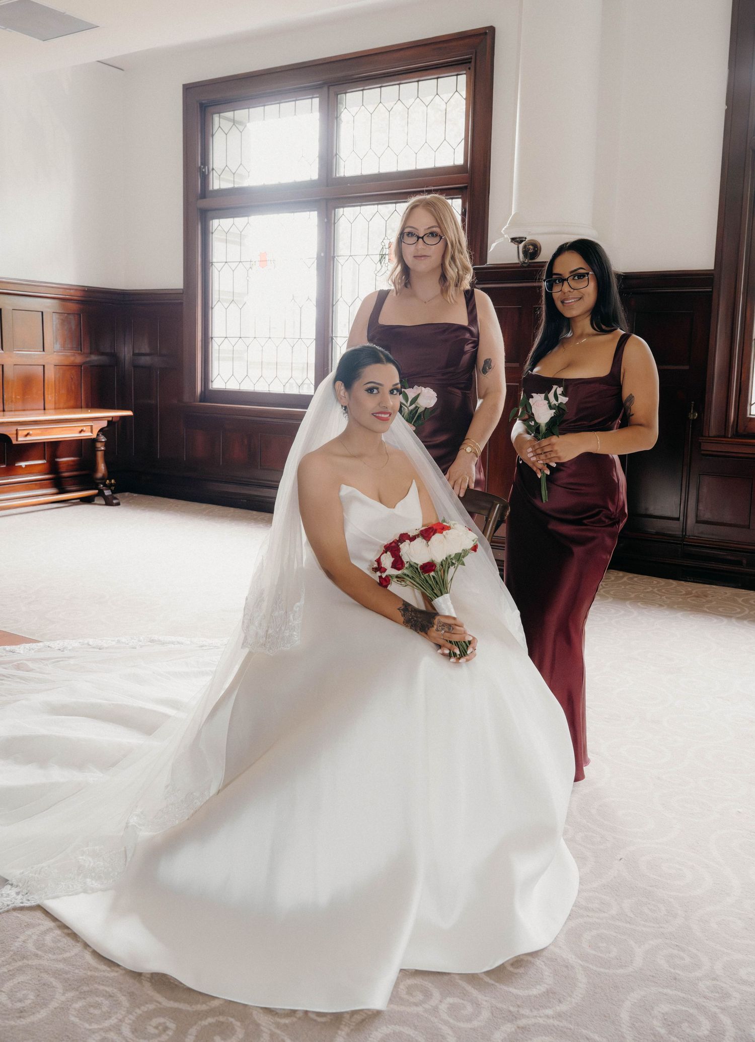 Bride in white ball gown with veil holds red bouquet flanked by two bridesmaids in burgundy satin dresses.